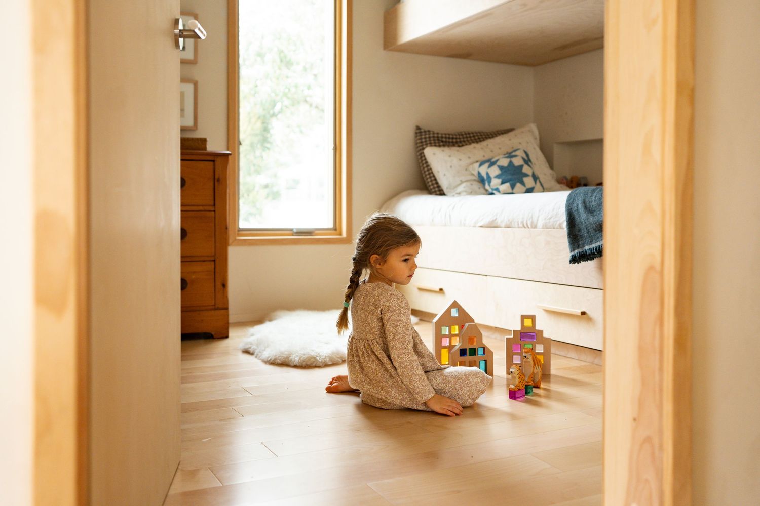 A child sits on a hardwood floor playing with toys near a cozy bed nook with natural window light.