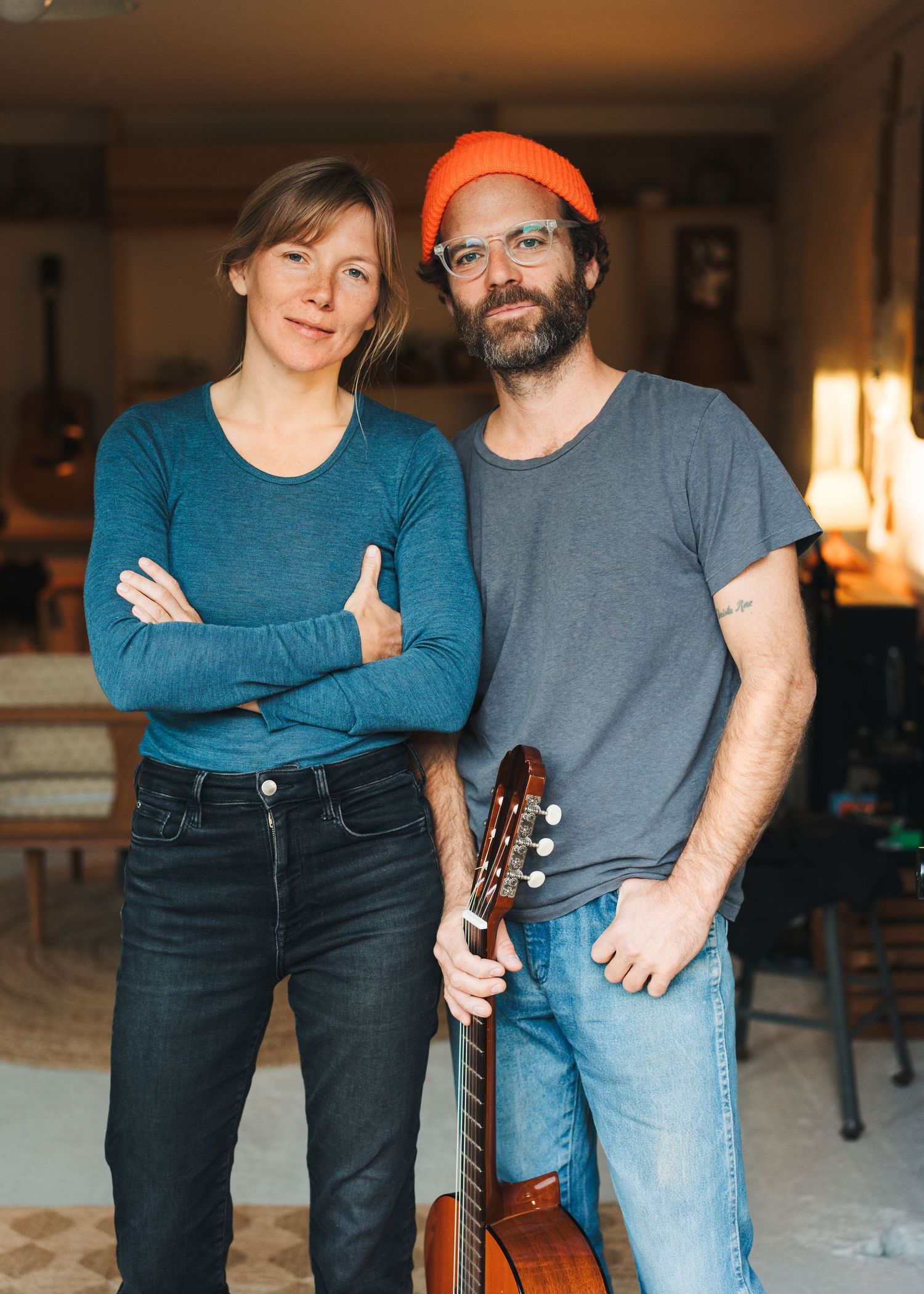 A couple in blue and gray casual wear poses with a guitar in a dimly lit indoor setting.
