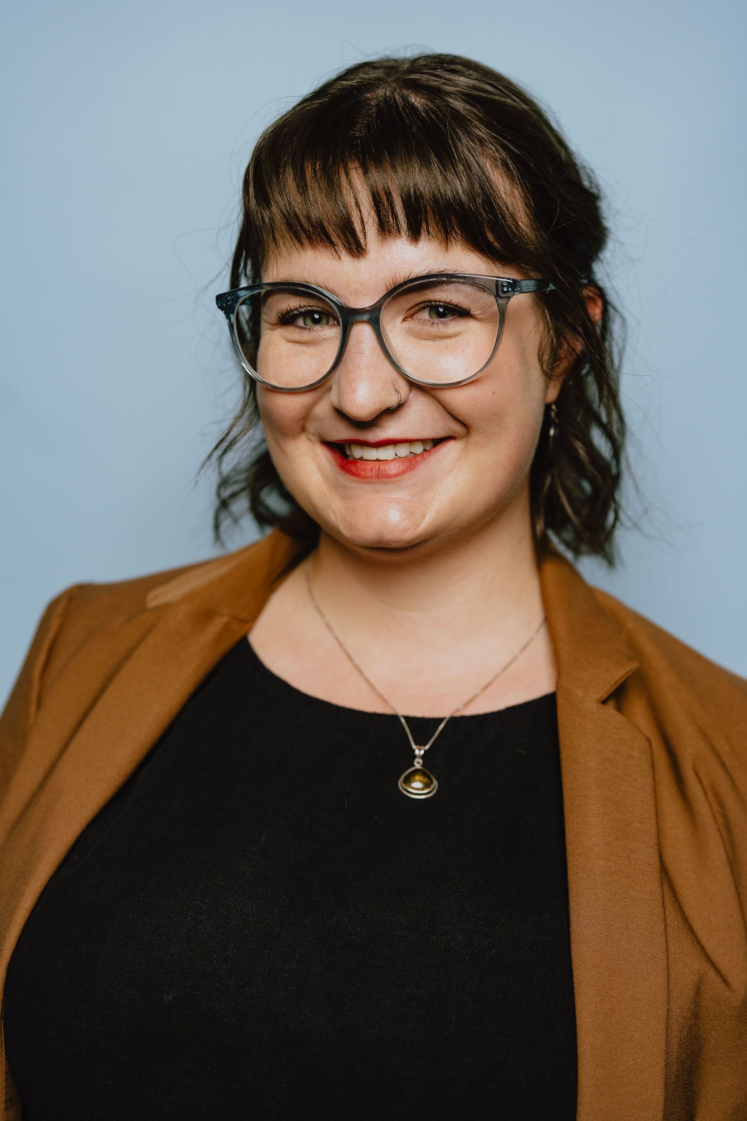 Professional headshot of someone wearing glasses, a black top and brown blazer against a light blue background.