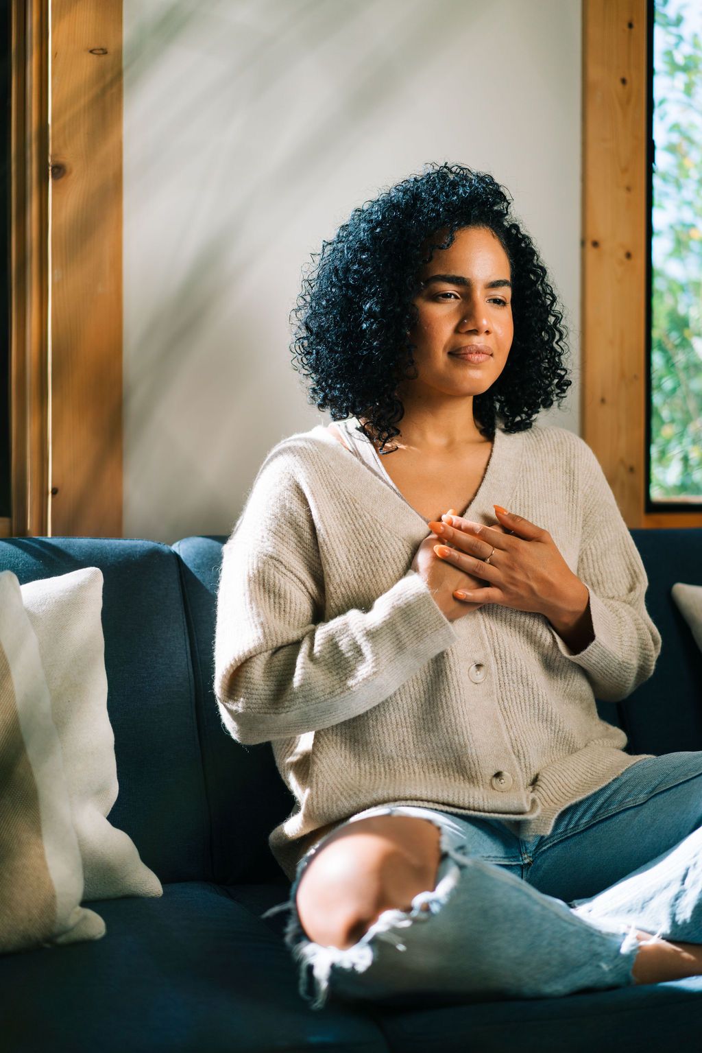 Person in white knit sweater relaxing on blue couch while using mobile phone in sunlit room.