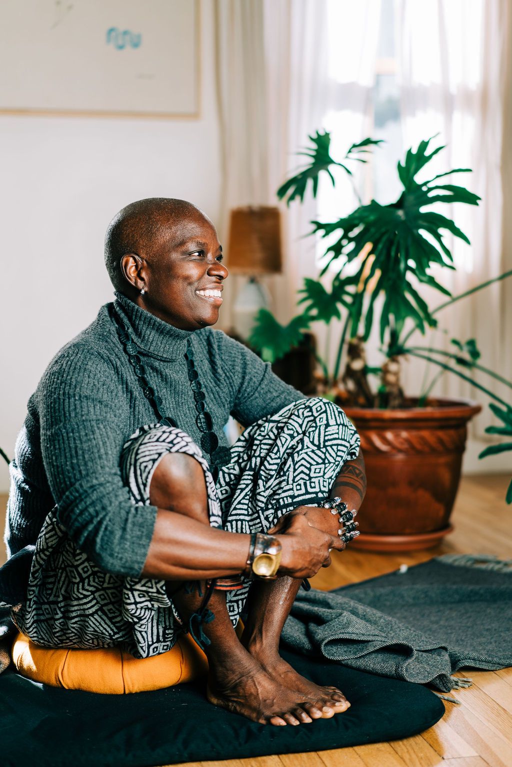 A person in a gray sweater relaxes on a floor cushion near houseplants in a bright, modern living space.