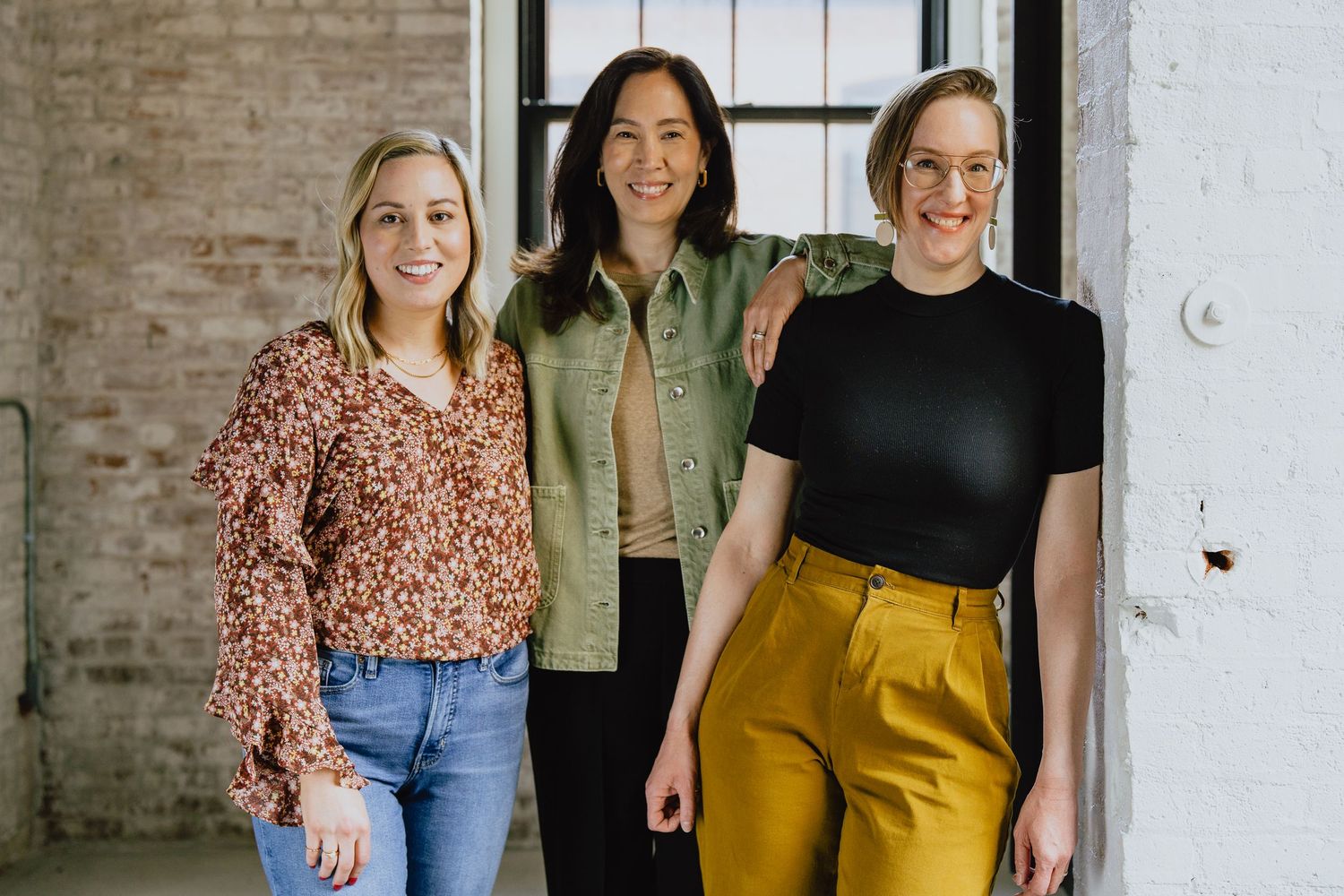A group of friends in casual attire standing together and smiling in front of a brick wall.