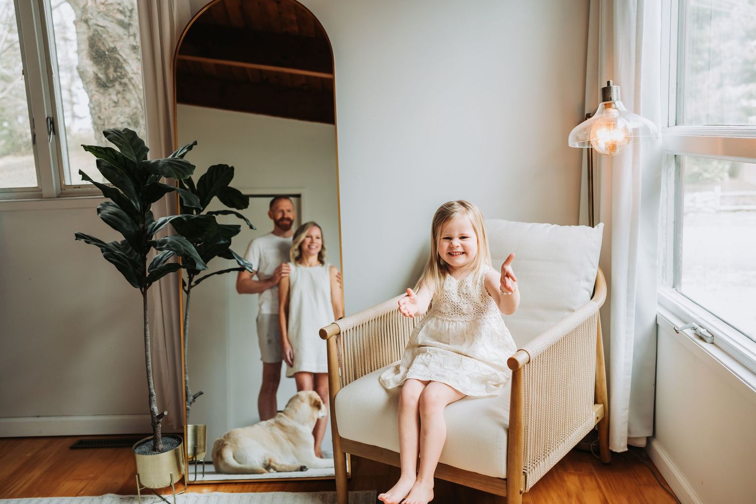 Young girl sitting in rattan chair smiling, with mother and dog reflected in large arched mirror.