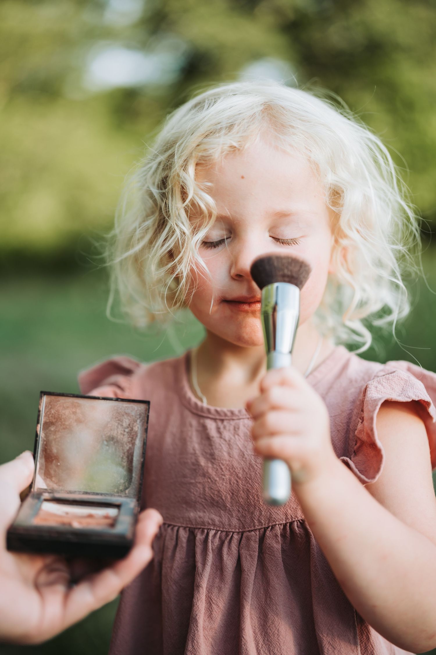 A young child in a pink dress plays with makeup, holding a brush and compact powder outdoors.