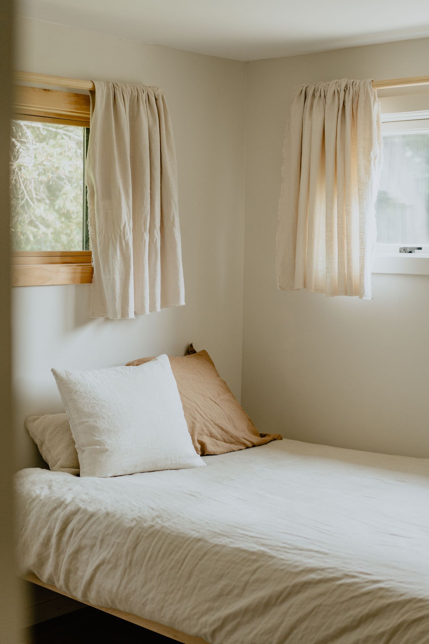 A serene minimalist bedroom interior with white linens and natural light filtering through beige curtains on small windows.