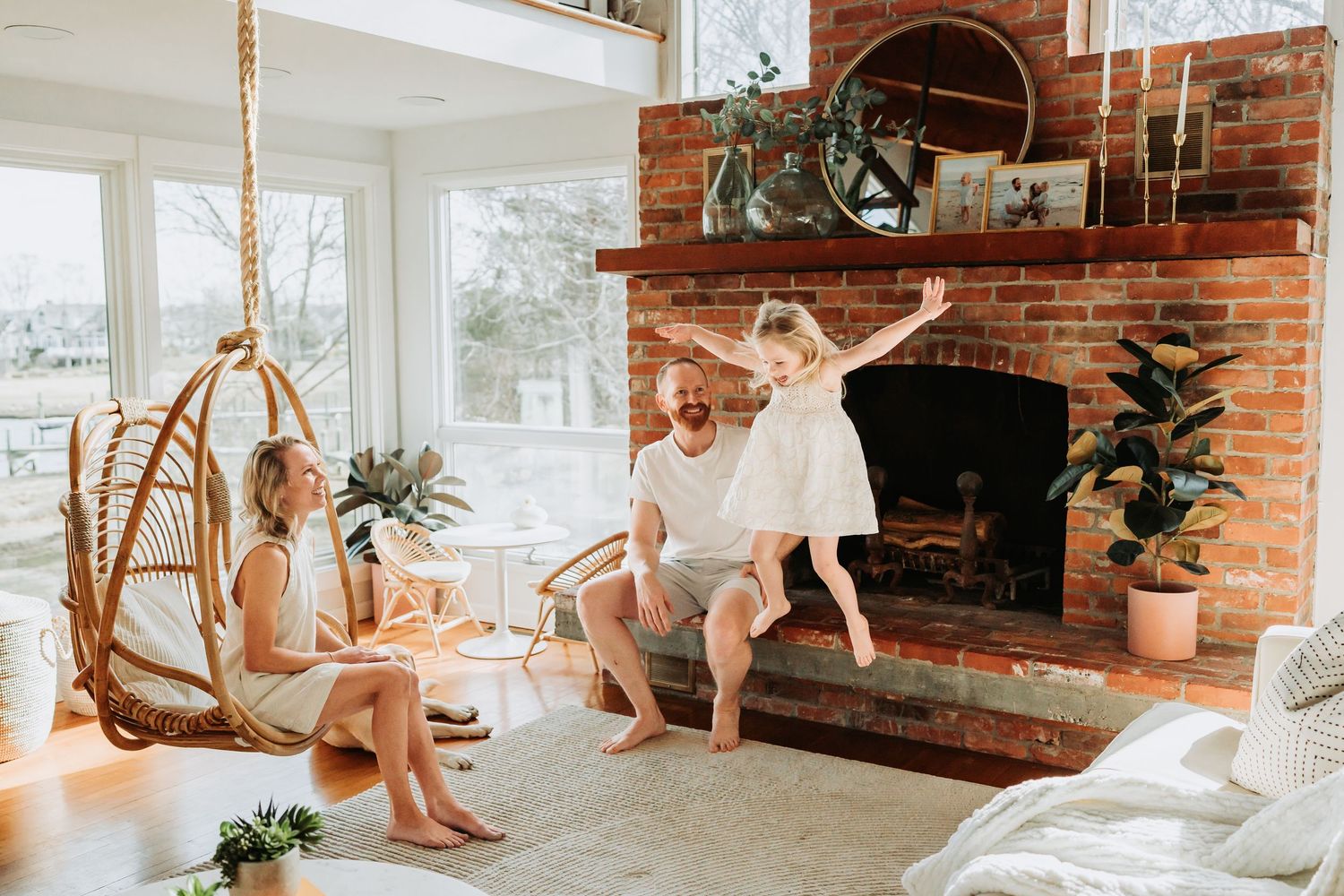 A joyful family plays by a brick fireplace in a bright living room with a rattan hanging chair.