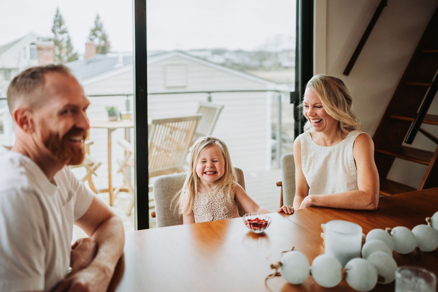 A joyful family laughs together at a dining table, with a blonde girl smiling between her parents near a large window.