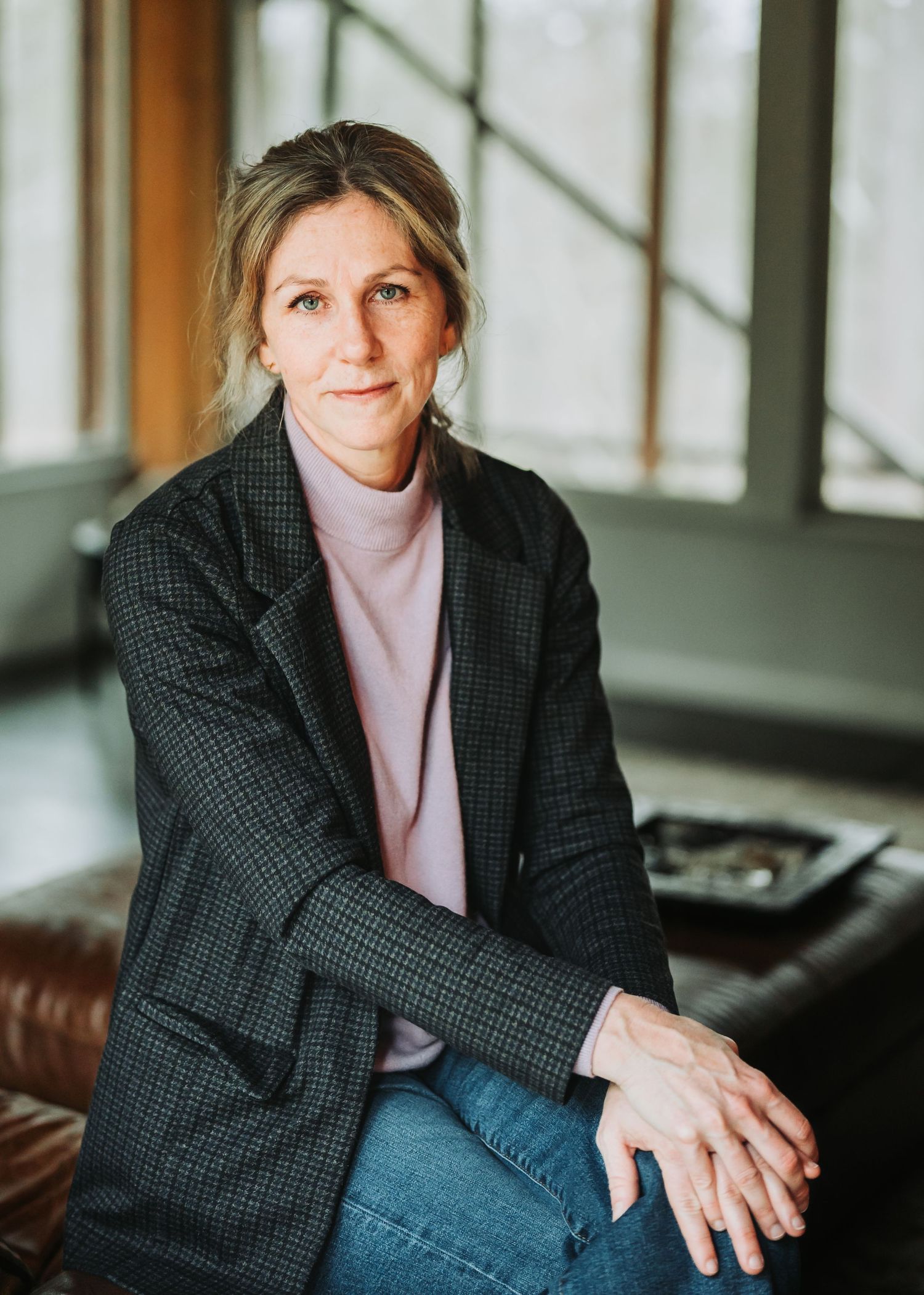 Professional in a dark checkered blazer and jeans sits casually on leather seating near bright windows.