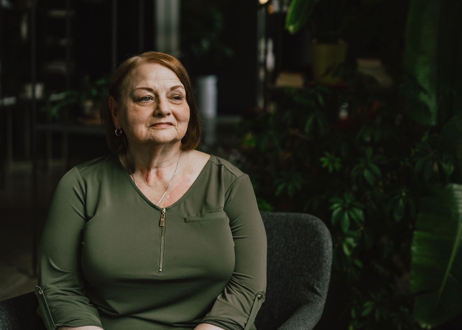 Portrait of person in olive green top seated in dark indoor setting with plants.