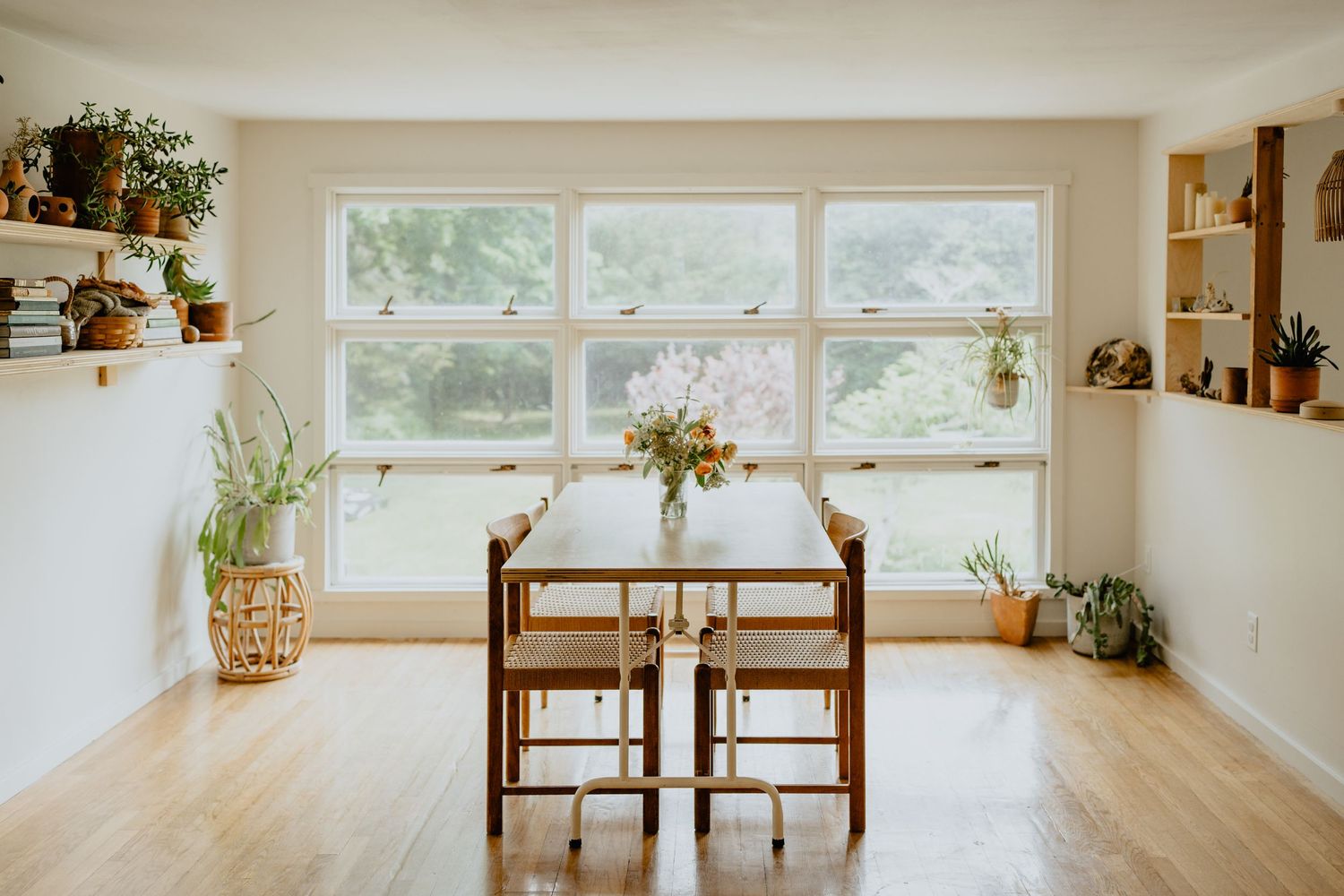 Bright minimalist dining area with wooden table, chairs, and plants on floating shelves near large windows.