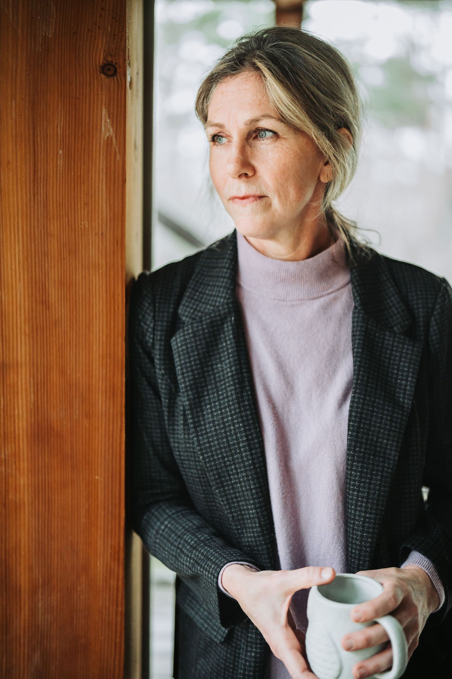 Person wearing grey blazer and light turtleneck holds white coffee cup while sitting near wooden panel wall.