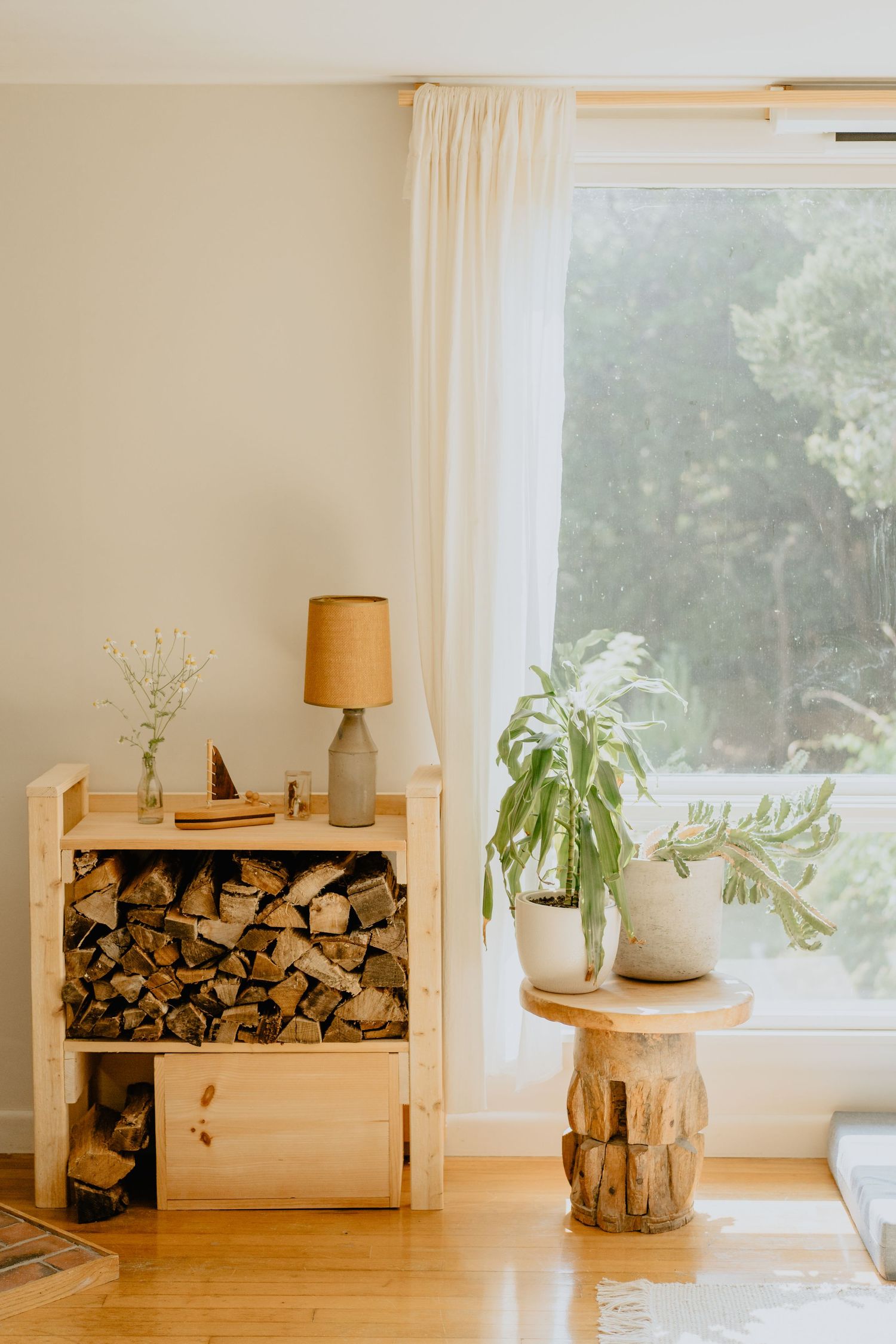 Rustic corner with firewood storage, table lamp, and potted plant by curtained window.