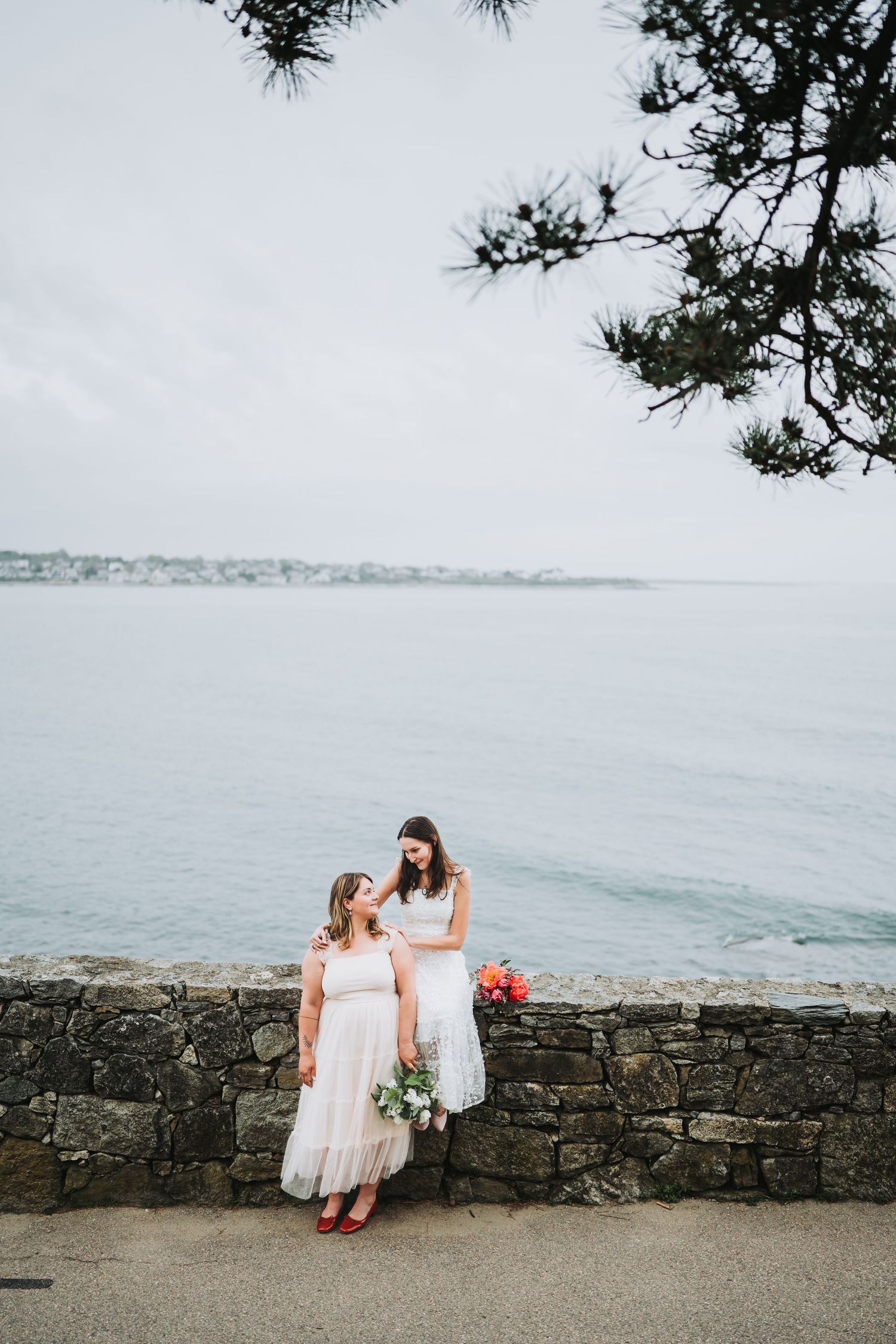 Two people in white dresses sit together on a stone wall overlooking the ocean on a cloudy day.