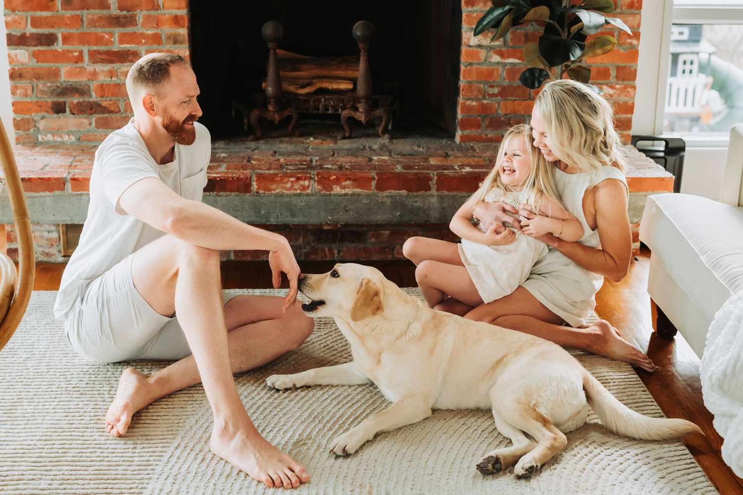 Family playing with yellow Labrador dog on rug by brick fireplace in cozy living room.