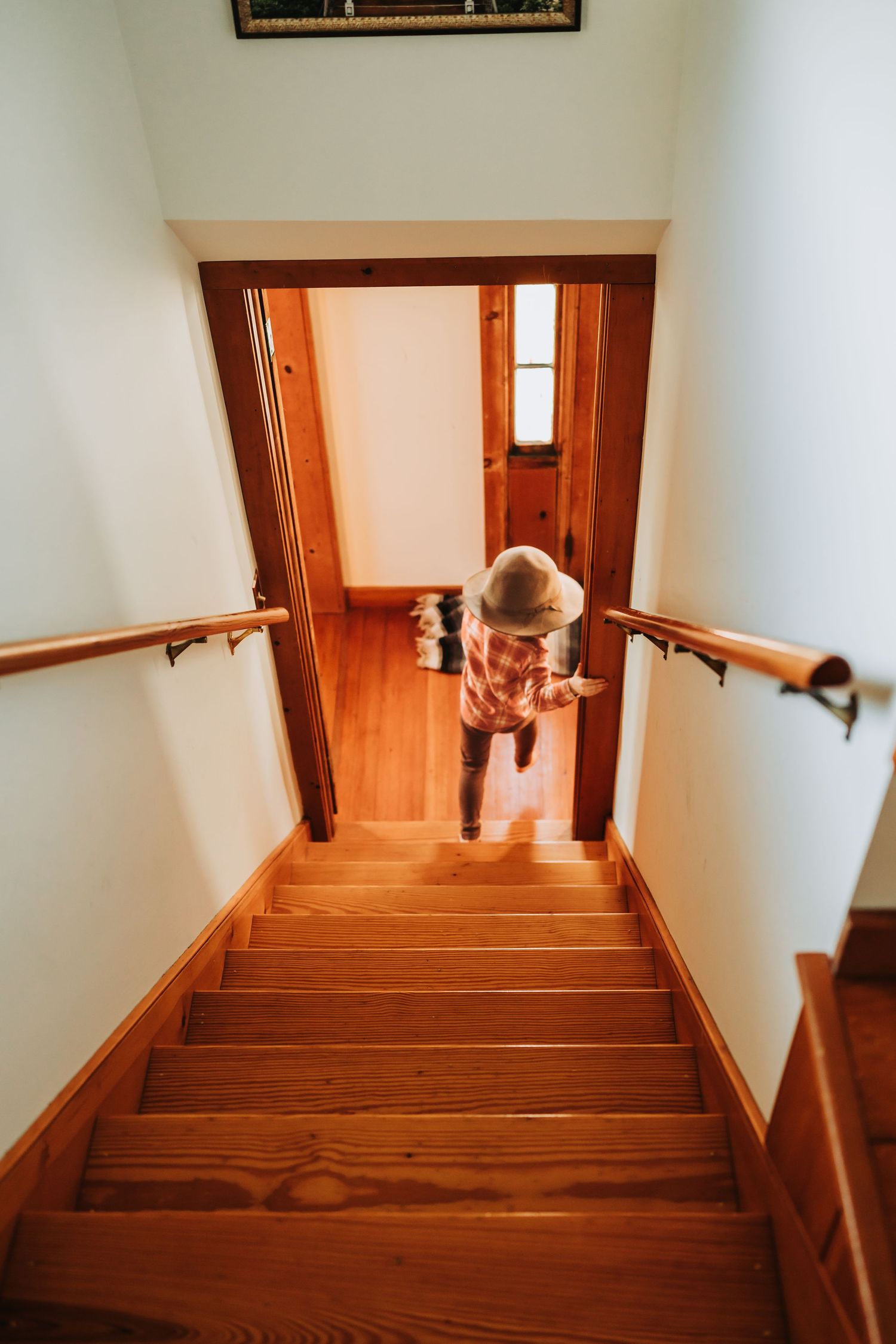 A dog peeks out from behind a wooden staircase in an indoor hallway with warm lighting and white walls.