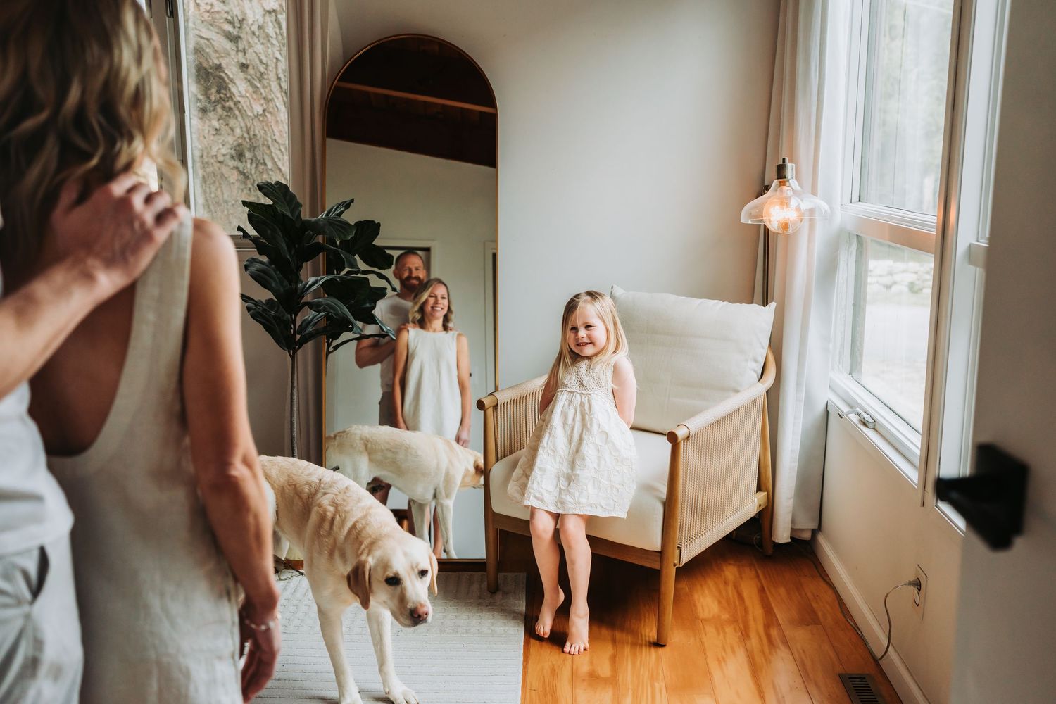 Little girl in white dress smiles near a window while a golden retriever walks beside a woman indoors.