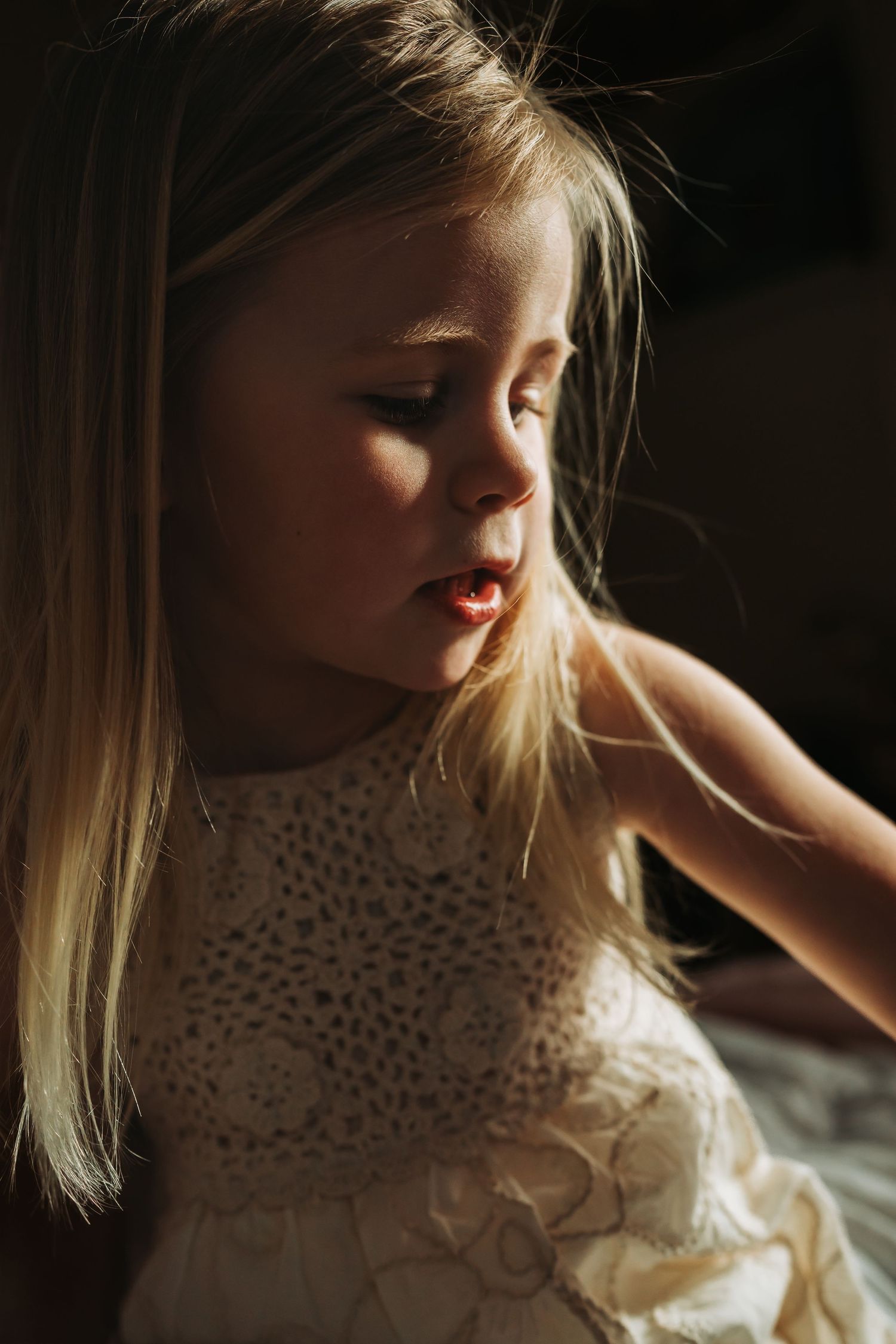 Young blonde girl in white lace dress with soft natural lighting casting warm shadows on her face.