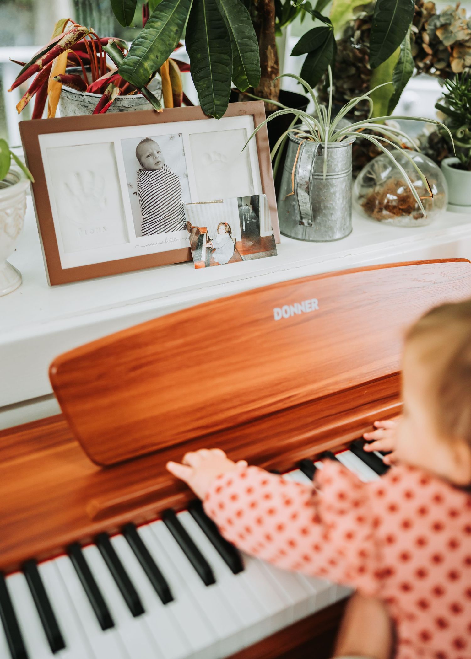 Small hands reaching across piano keys with polka dot fabric and indoor plants visible in background.