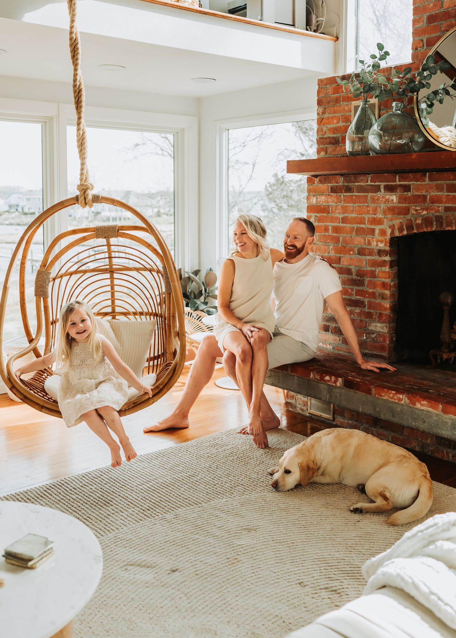 Family with young girl in hanging rattan chair beside brick fireplace with yellow lab dog in bright living room.