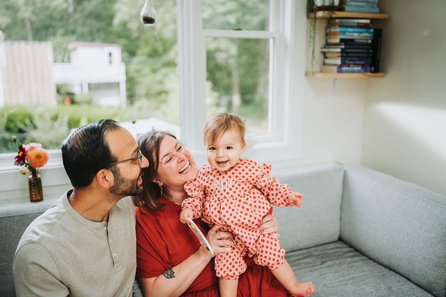 A joyful family moment shared on a gray couch near a bright window with a bookshelf in the background.