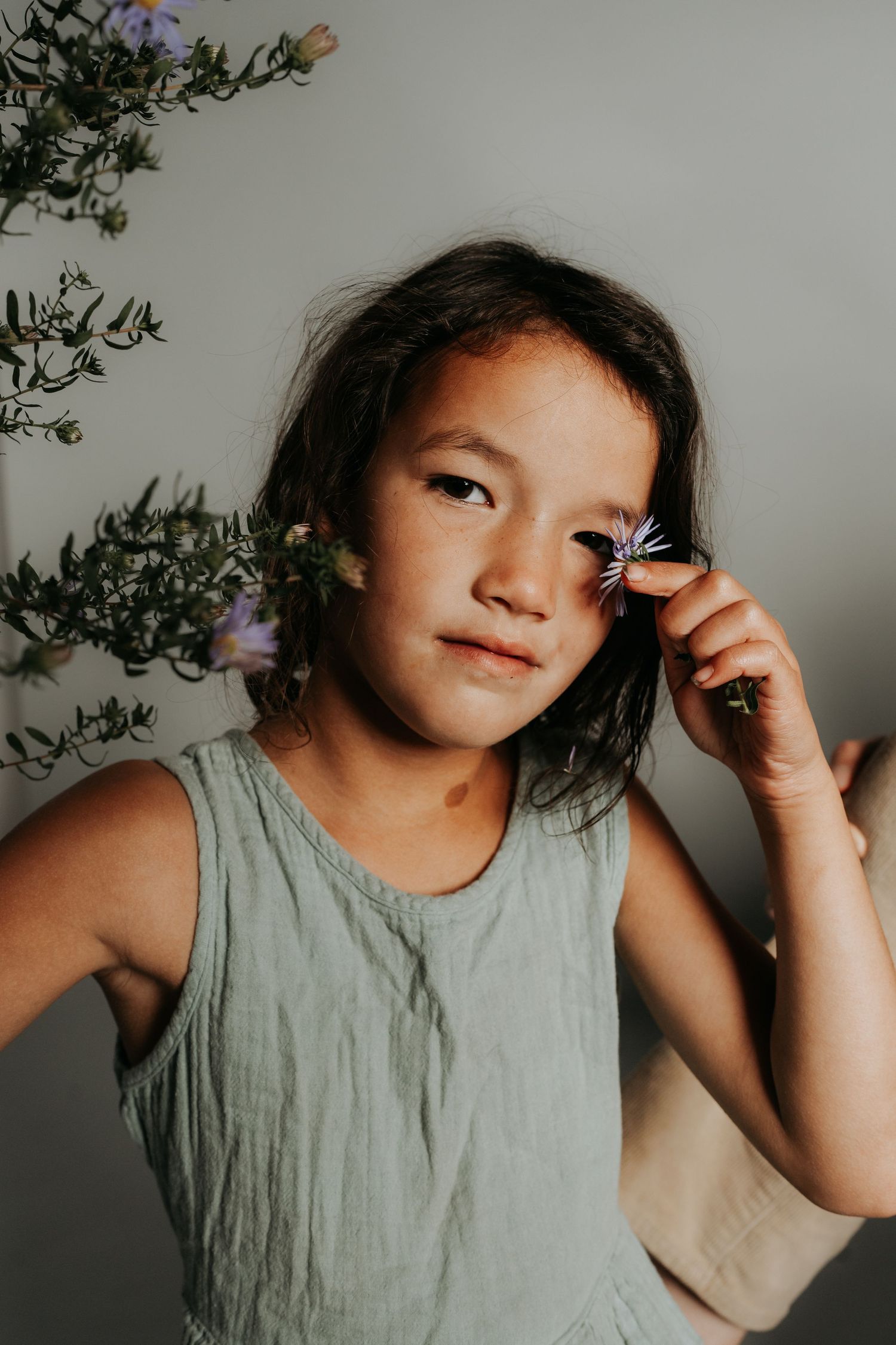 Young child in gray tank top poses with flowers against neutral background.