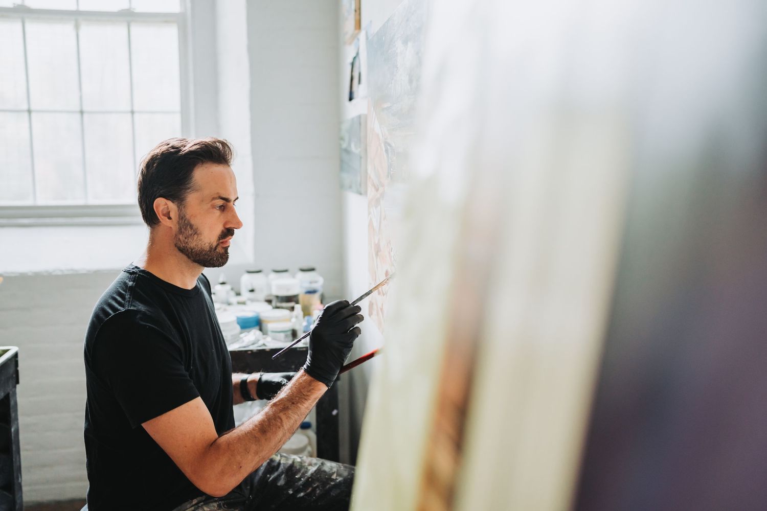 Artist in black t-shirt painting at easel by bright window in sunlit studio space.