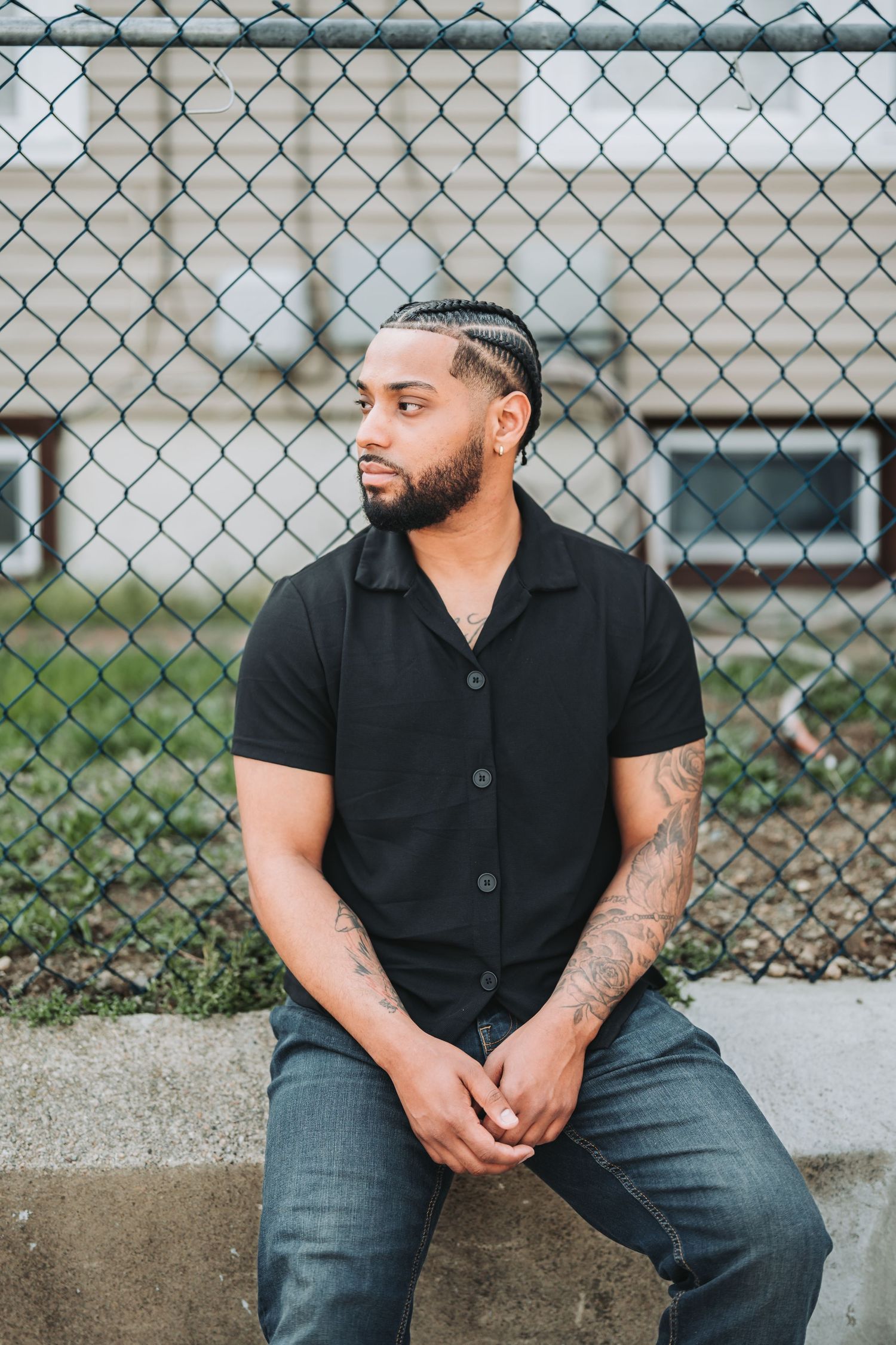 Person in black polo shirt and gray pants sits thoughtfully by chain link fence in outdoor setting.