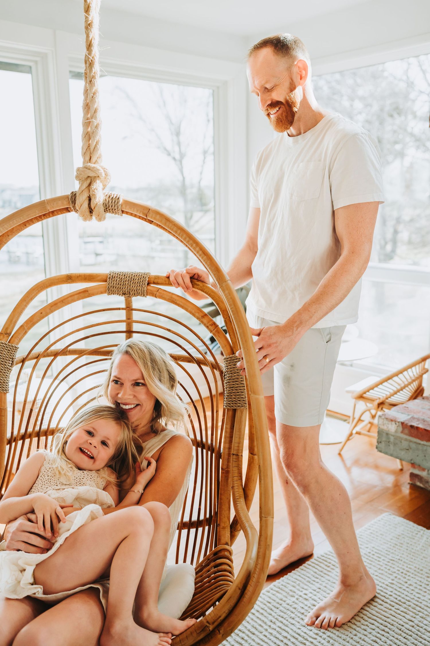 Happy family with young daughter relaxing in a rattan hanging chair in a bright sunroom.