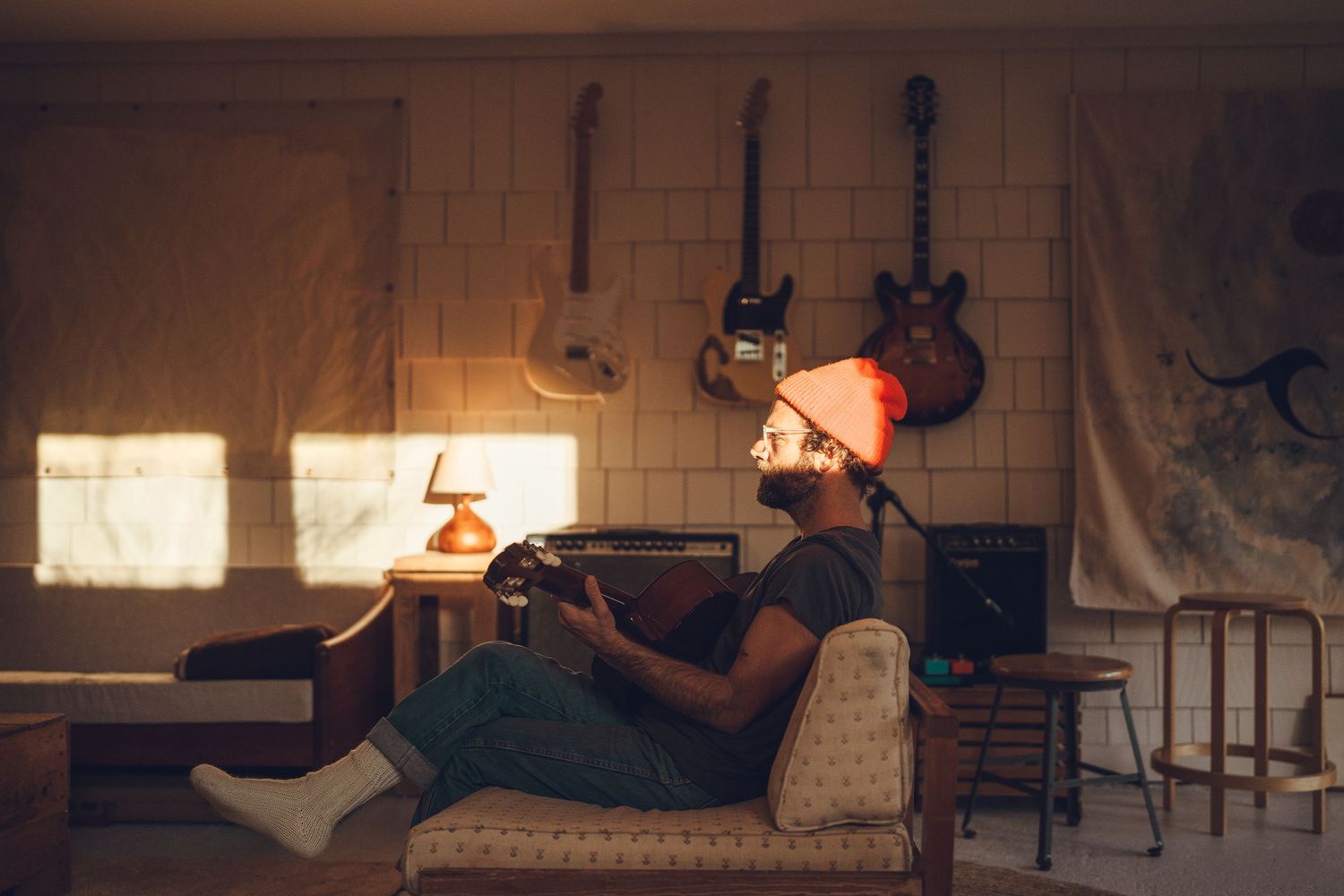 A person reclines on a vintage couch reading in warm sunlight while musical instruments hang on the wall behind them.