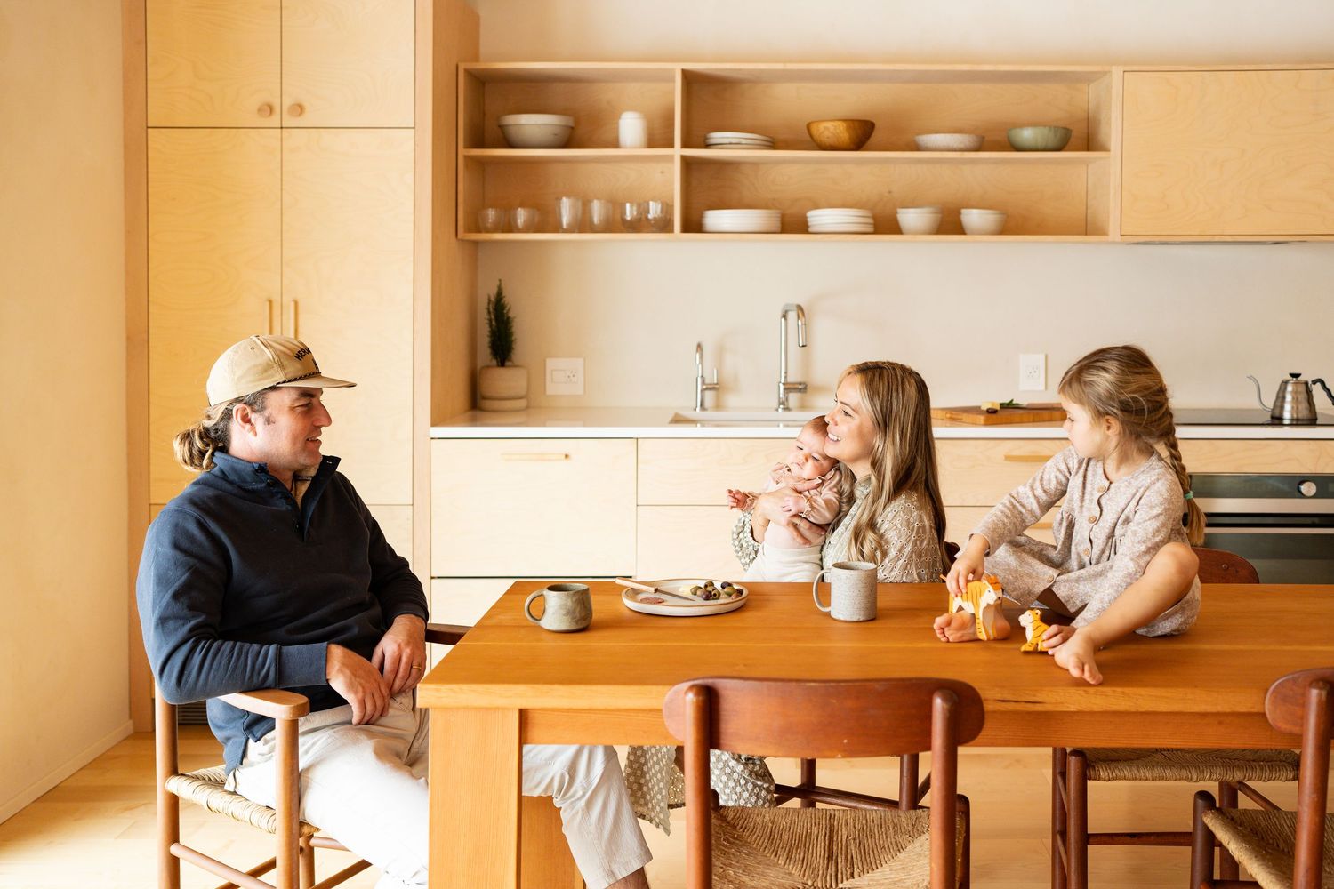 Family members gather around a rustic wooden dining table in a warmly lit kitchen with natural decor.