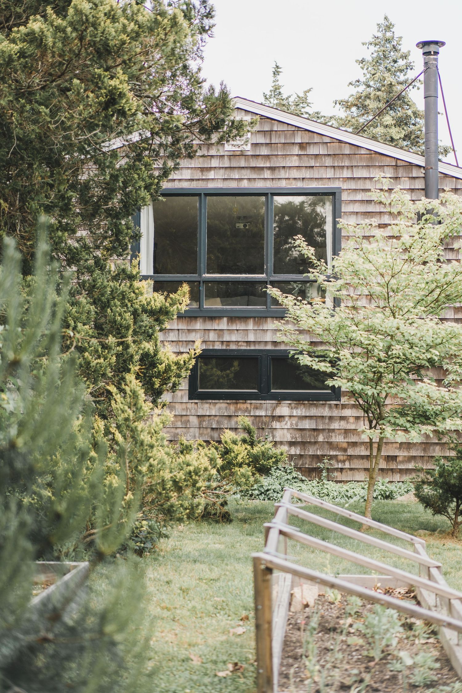 A modern ranch-style house with cedar shingle siding and large windows sits nestled among green trees and foliage.