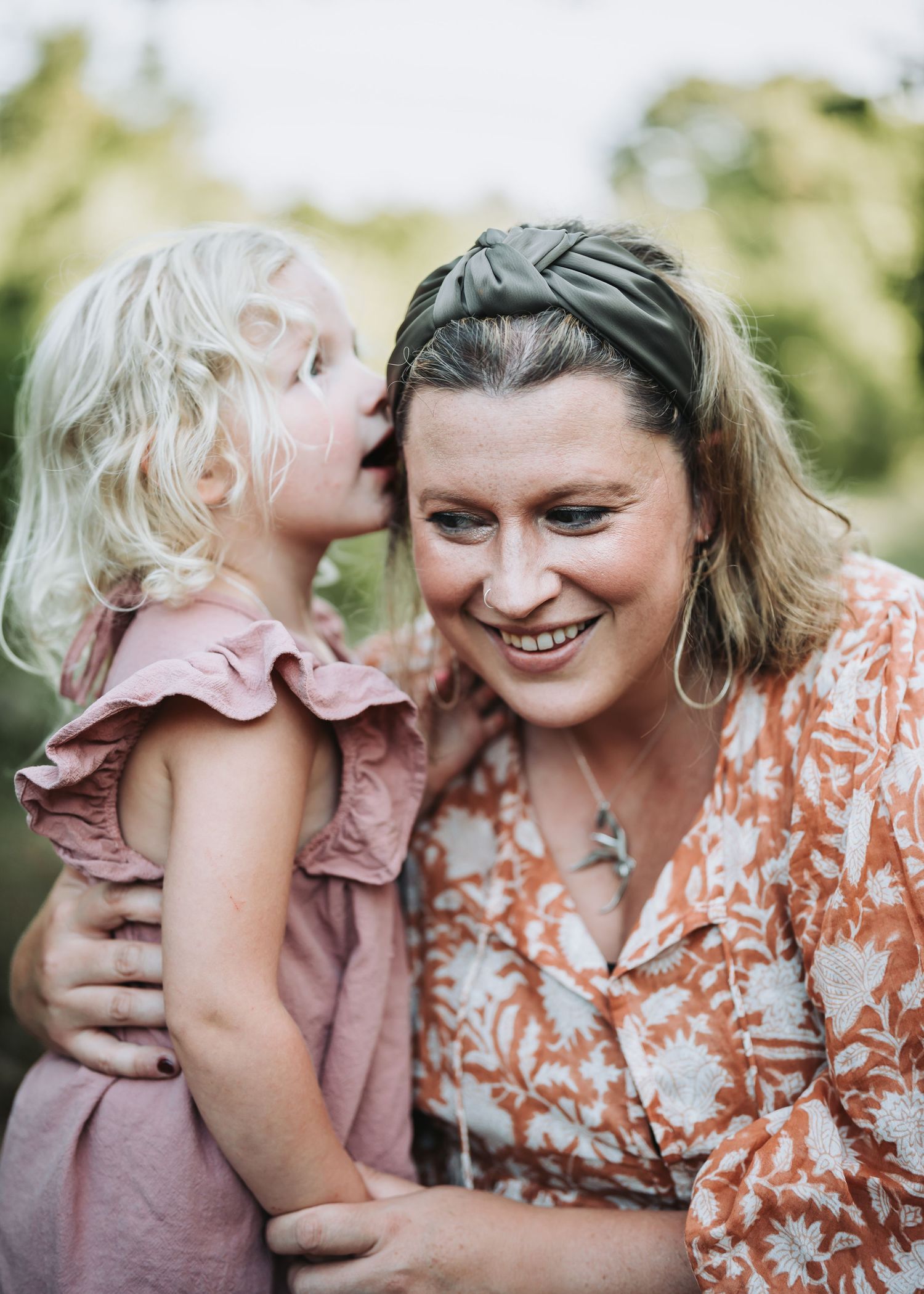 A sweet moment captured outdoors as child whispers to woman wearing floral print and headband.