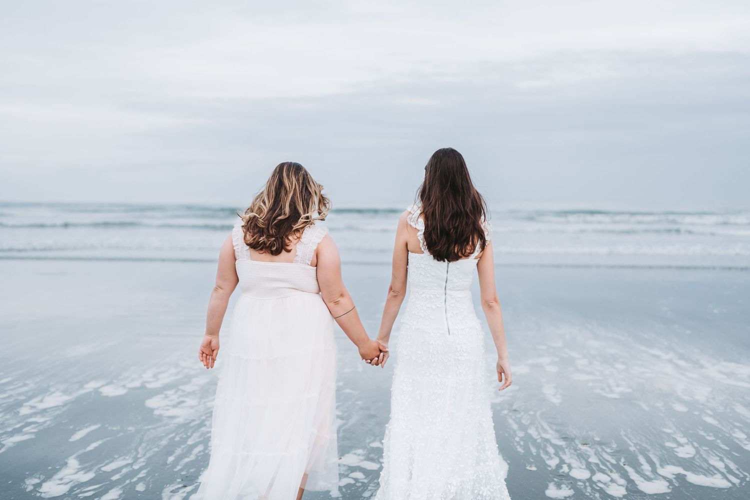 Two people in white dresses walking along a misty beach shore with waves in the background.