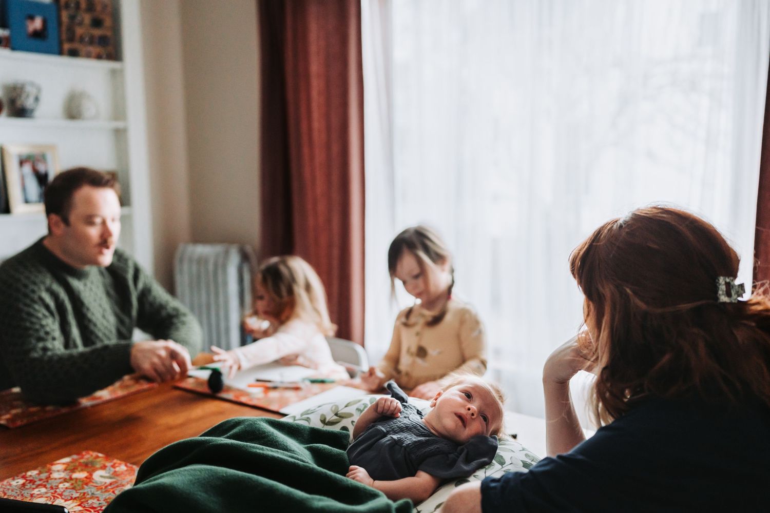 Family gathering around a wooden table with a baby wrapped in a green blanket.