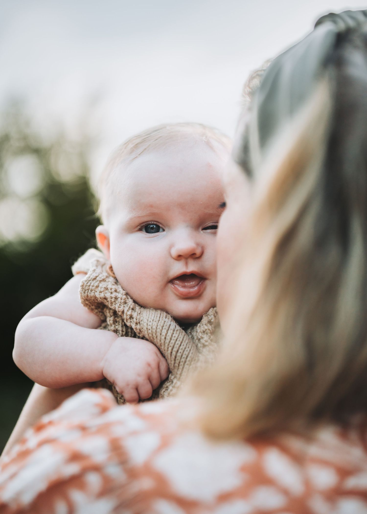 Close up of infant wearing beige knit outfit against outdoor background.