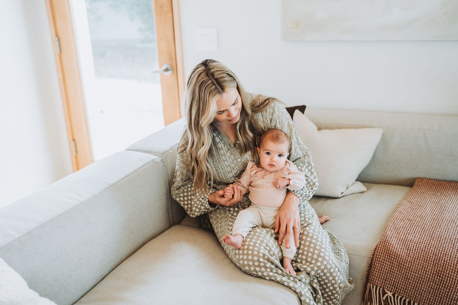 A mother in a cozy striped sweater holds her baby in a bright, minimalist living room setting.
