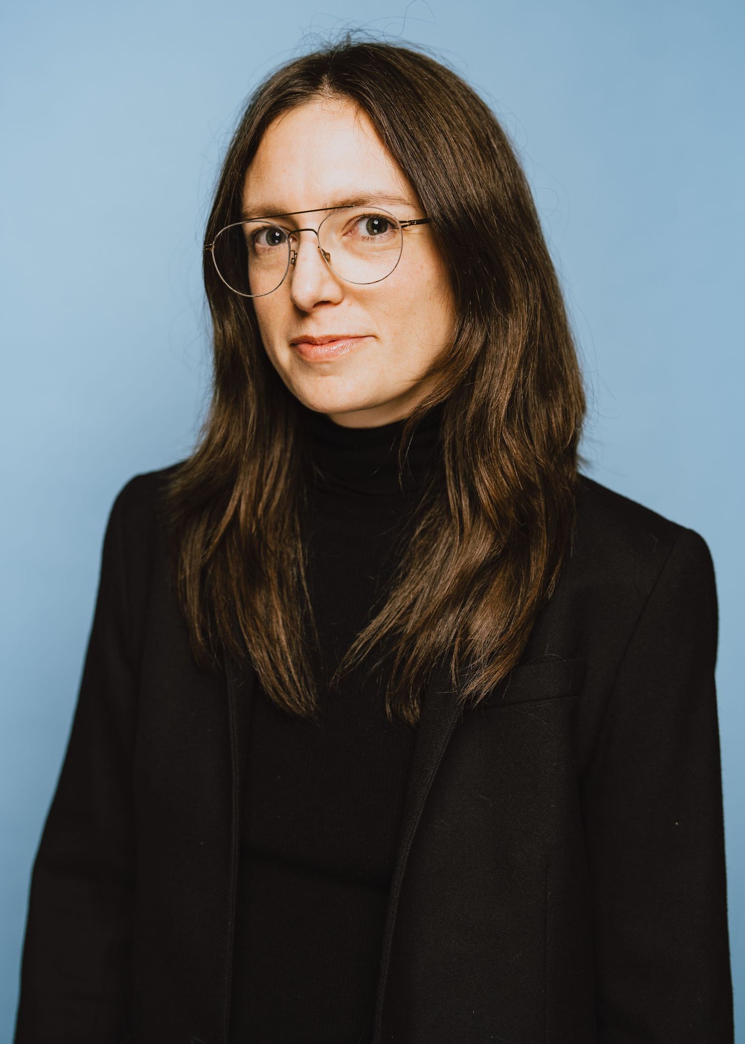 Professional portrait of someone wearing glasses, a black turtleneck and blazer against a light blue background.