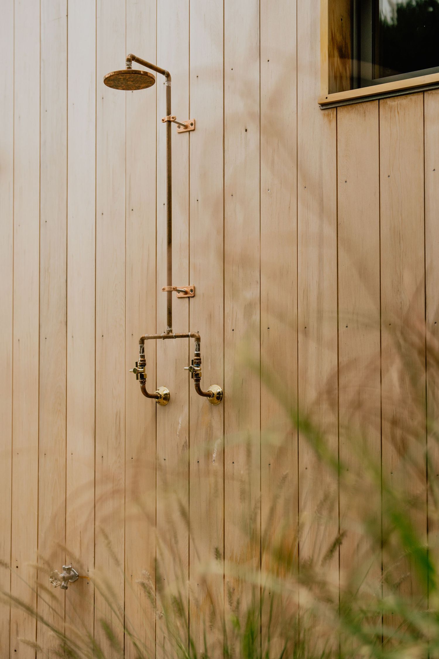 Modern outdoor shower with brass fixtures mounted on natural wood siding against beige wall.