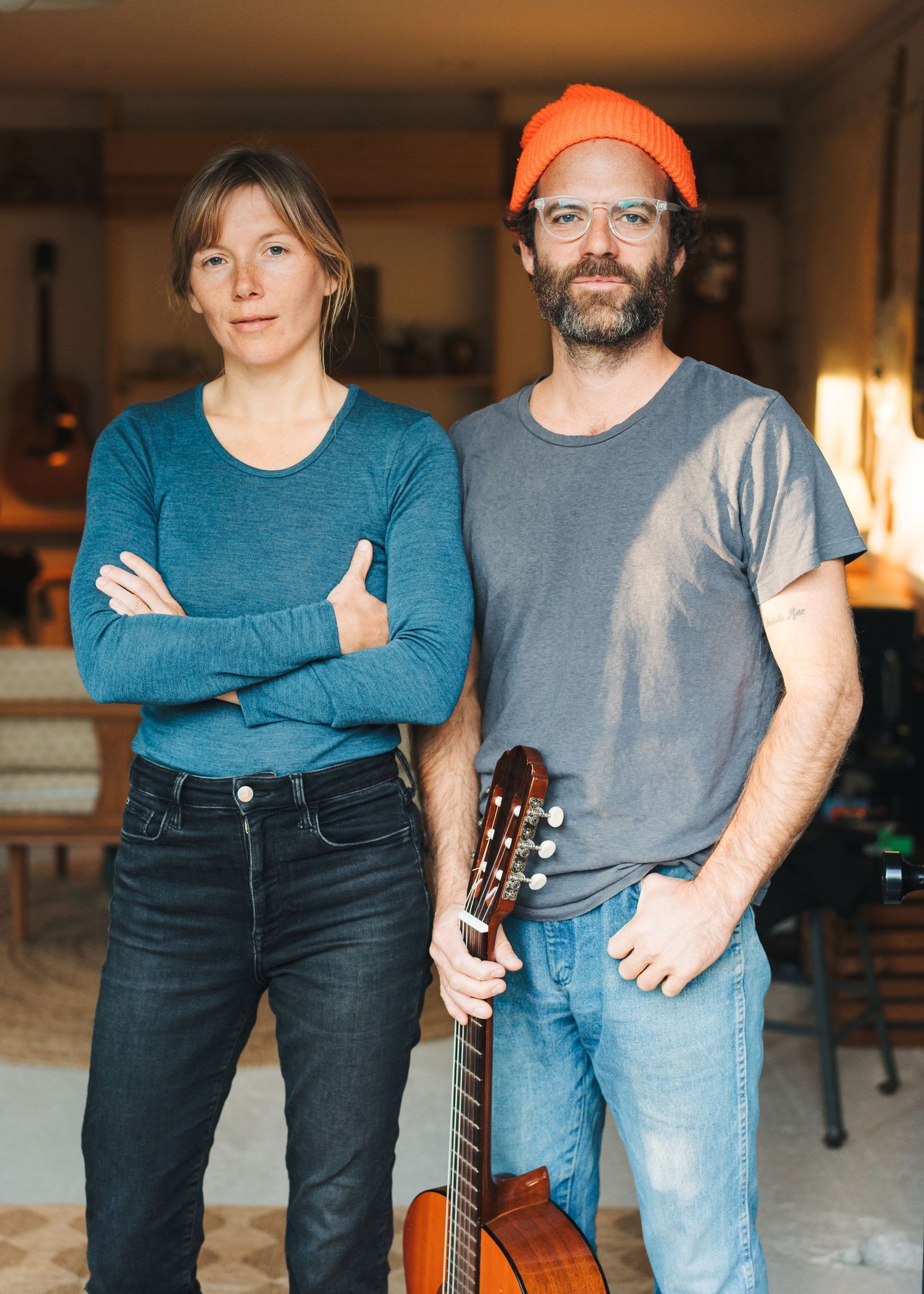 Two musicians pose indoors, one wearing an orange beanie and holding an acoustic guitar.