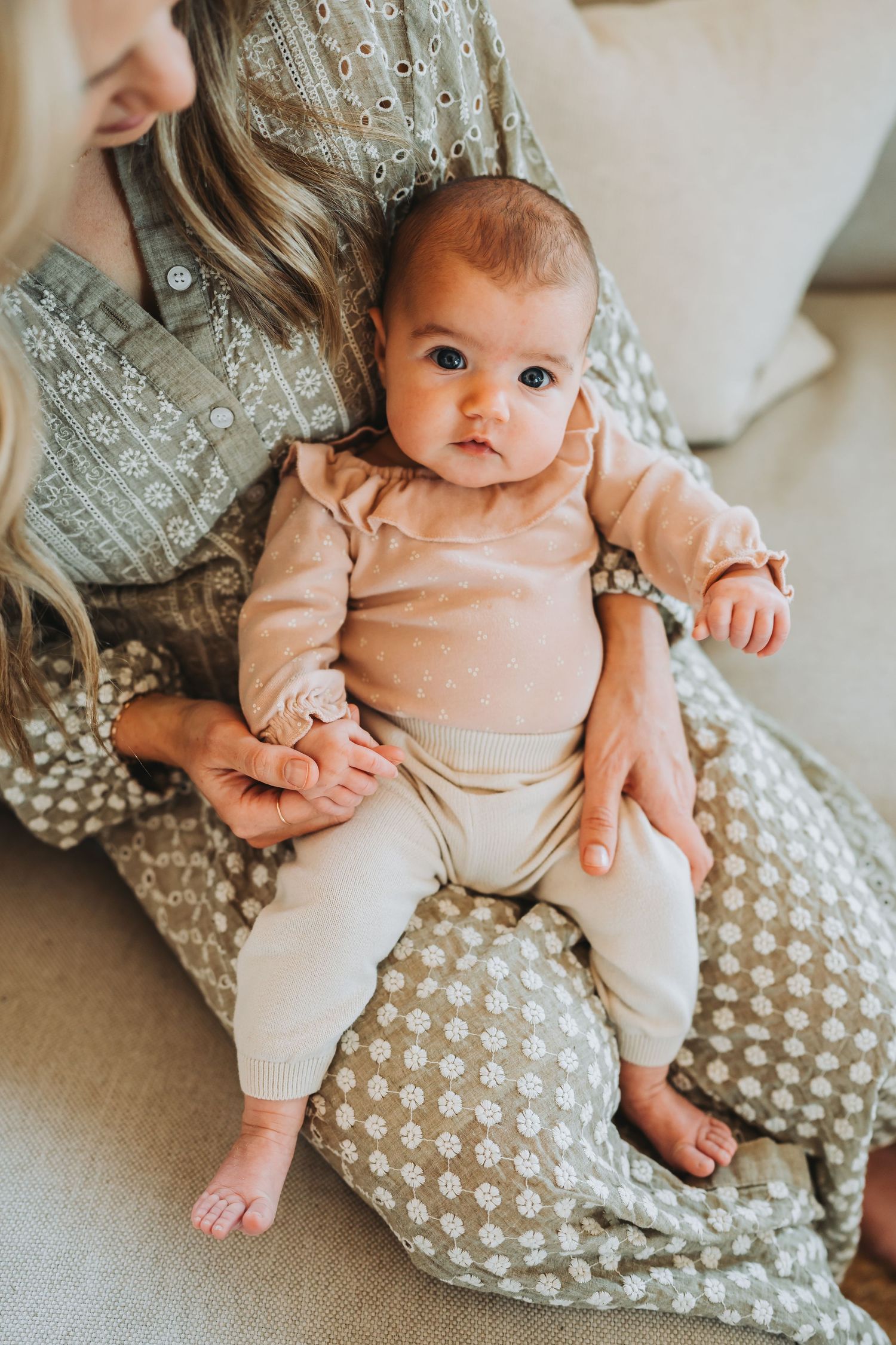Baby wearing peach outfit sits on patterned pillow in bright interior setting.