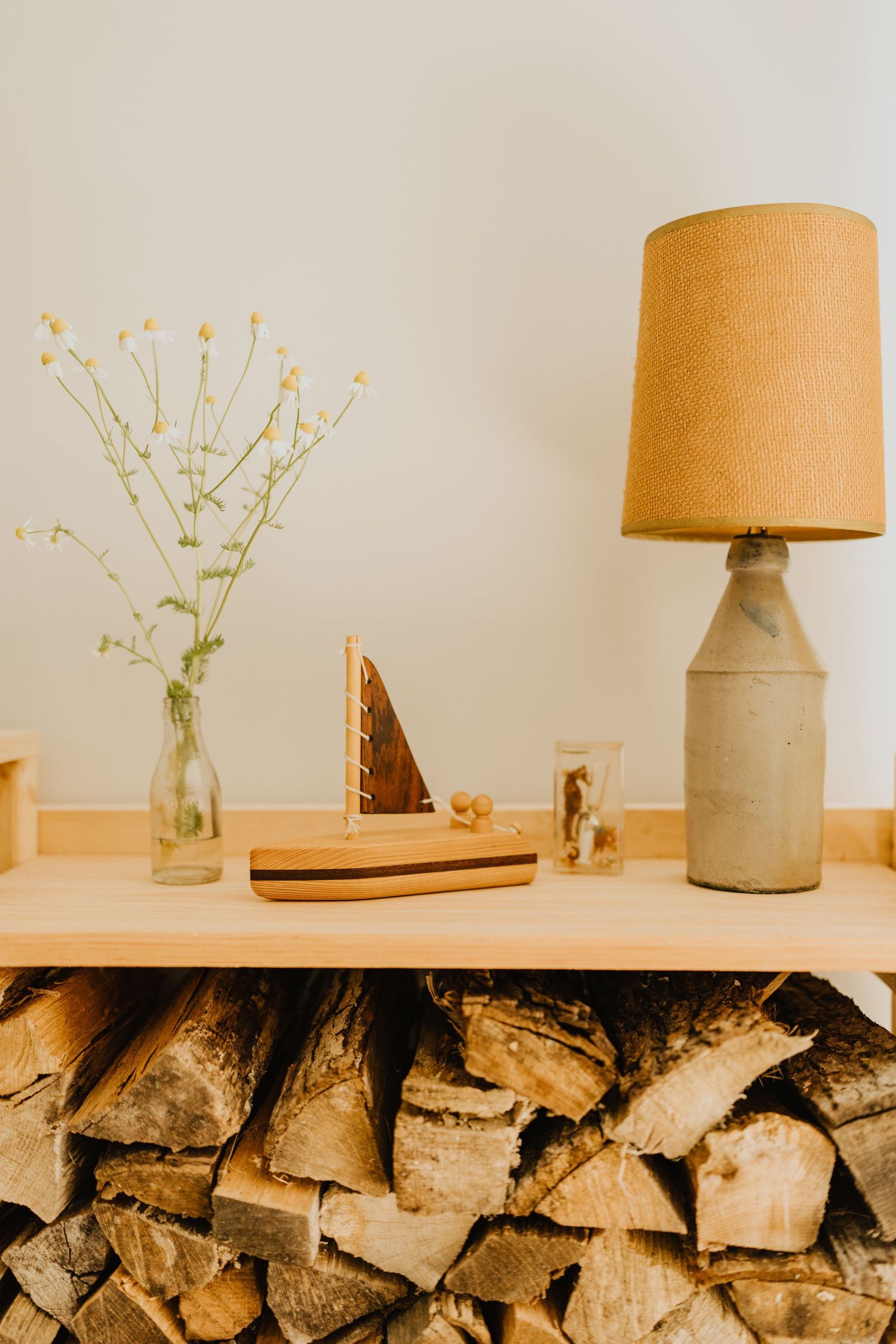 Modern concrete lamp and delicate flowers in glass vases displayed on shelf above stacked firewood.
