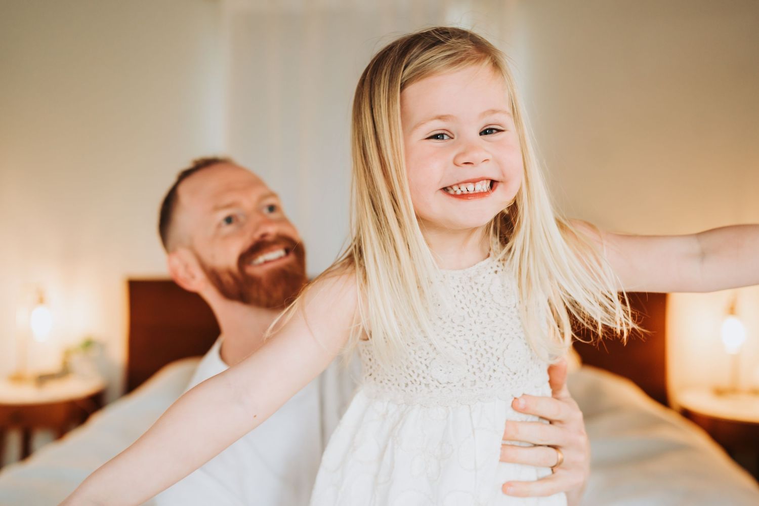 Happy little girl in white dress laughing while father plays with her in a warmly lit bedroom.