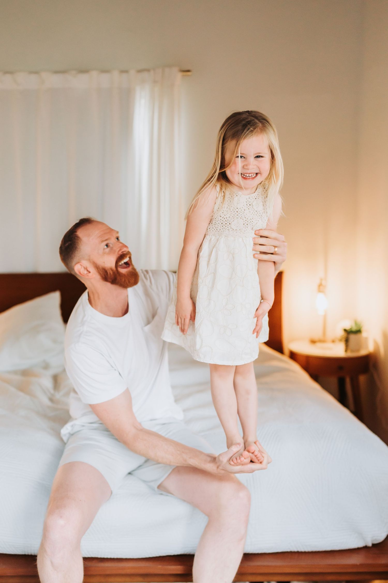 Laughing father in white outfit lifting happy toddler girl standing on bed in bright bedroom.