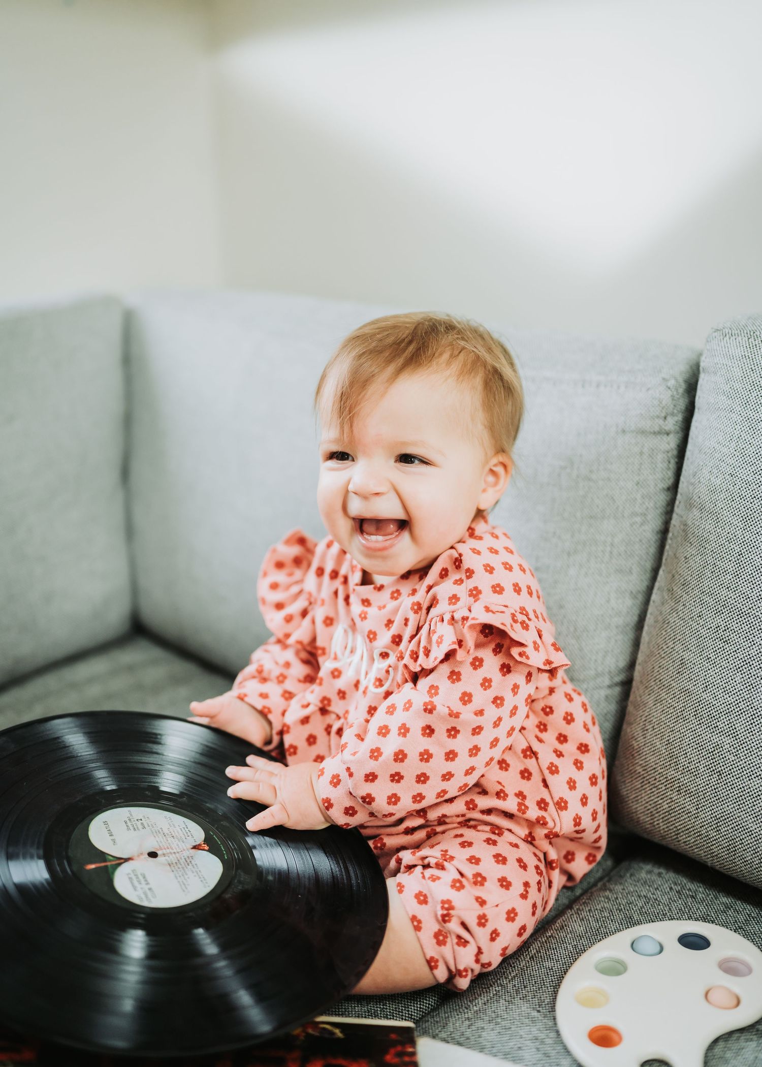 Baby in polka dot pajamas sits on couch laughing while playing with vinyl record.