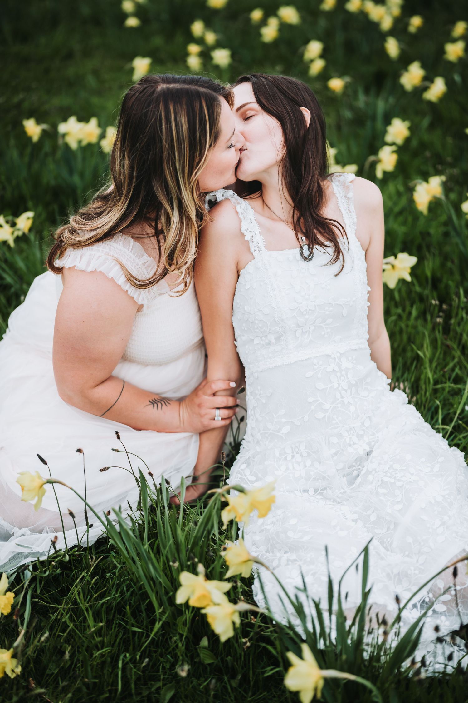 Two people in white lace dresses share an intimate moment while sitting in a field of yellow daffodils.