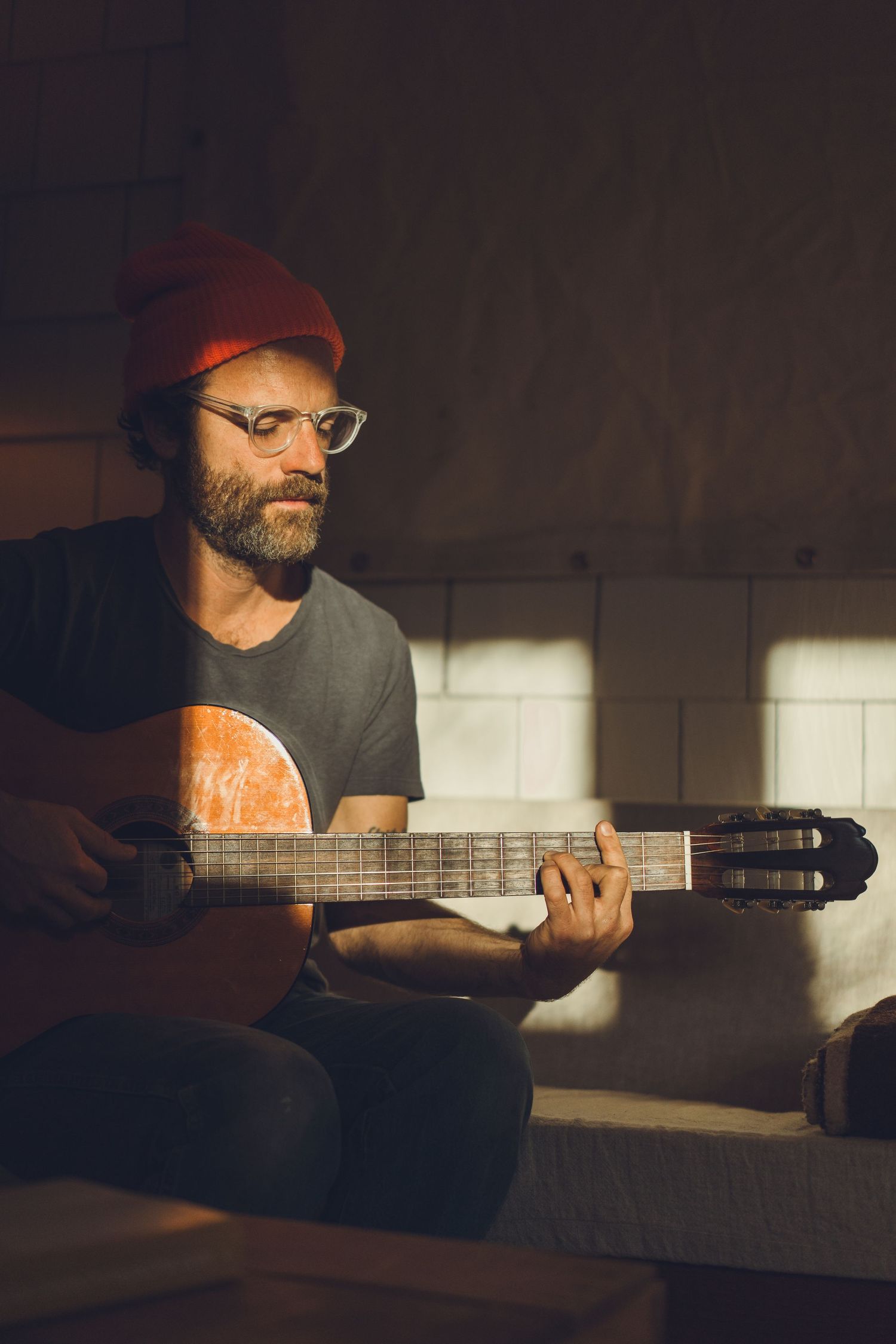 Musician in orange beanie plays acoustic guitar in moody indoor lighting.