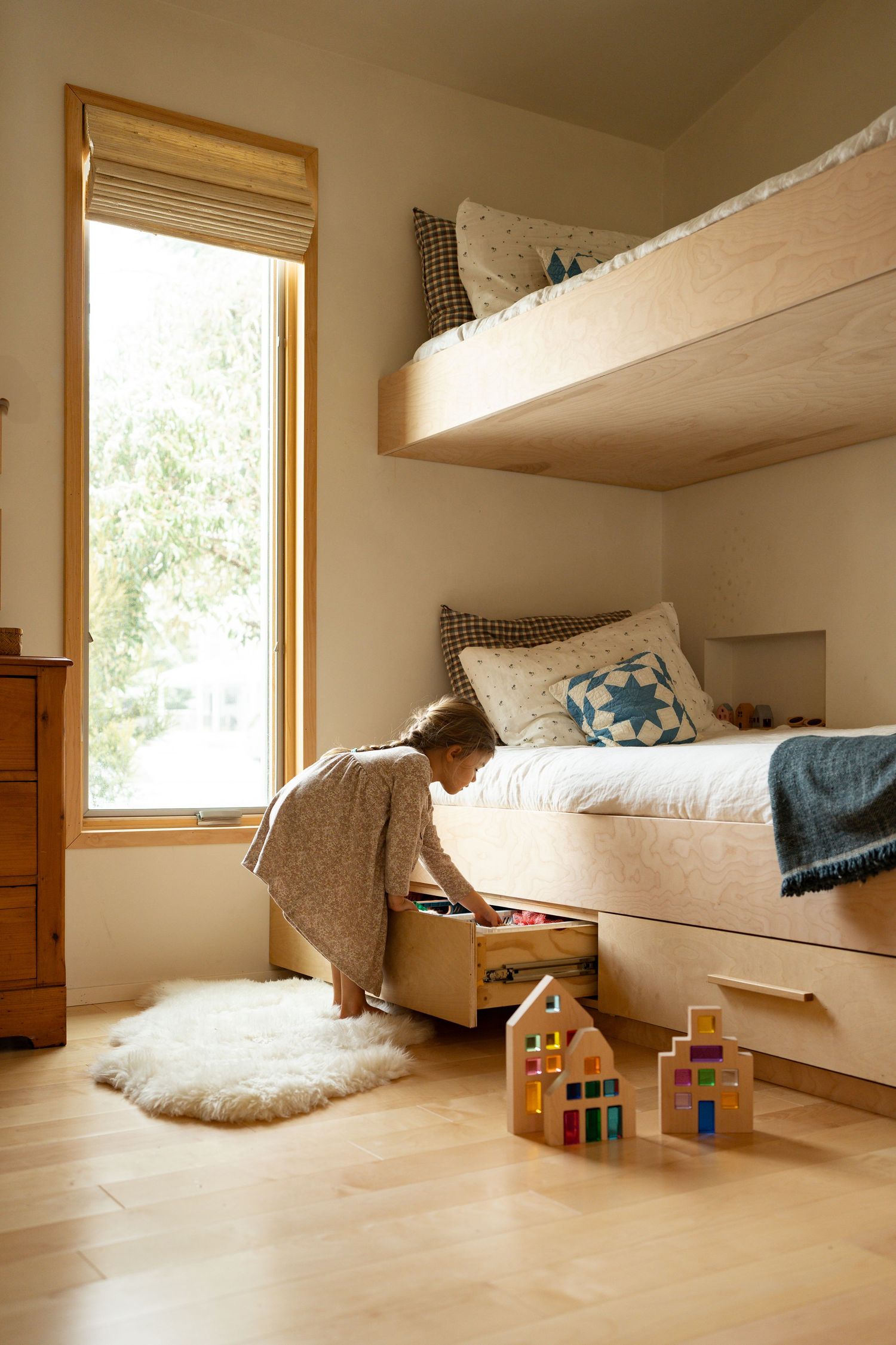 A cozy loft space with built-in bunk beds and natural light streaming through windows, featuring a small wooden dollhouse.