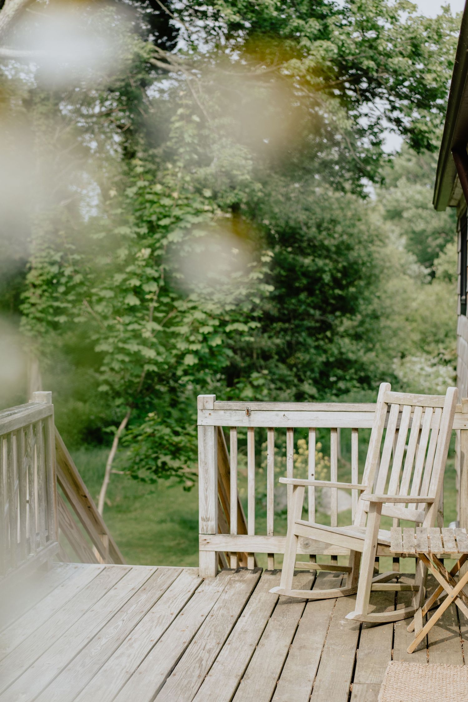 Weathered wooden rocking chairs line a rustic porch deck overlooking green foliage.