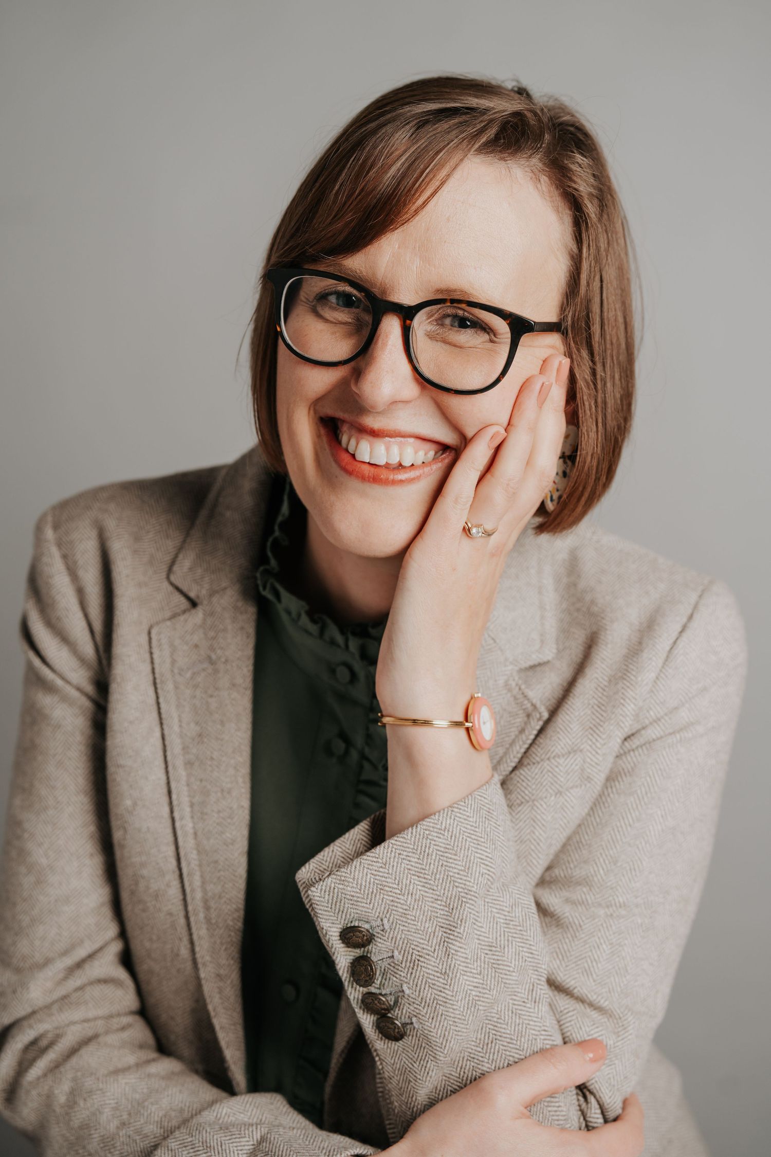 Professional headshots showing joyful expressions in a beige blazer and dark top against a neutral gray background.