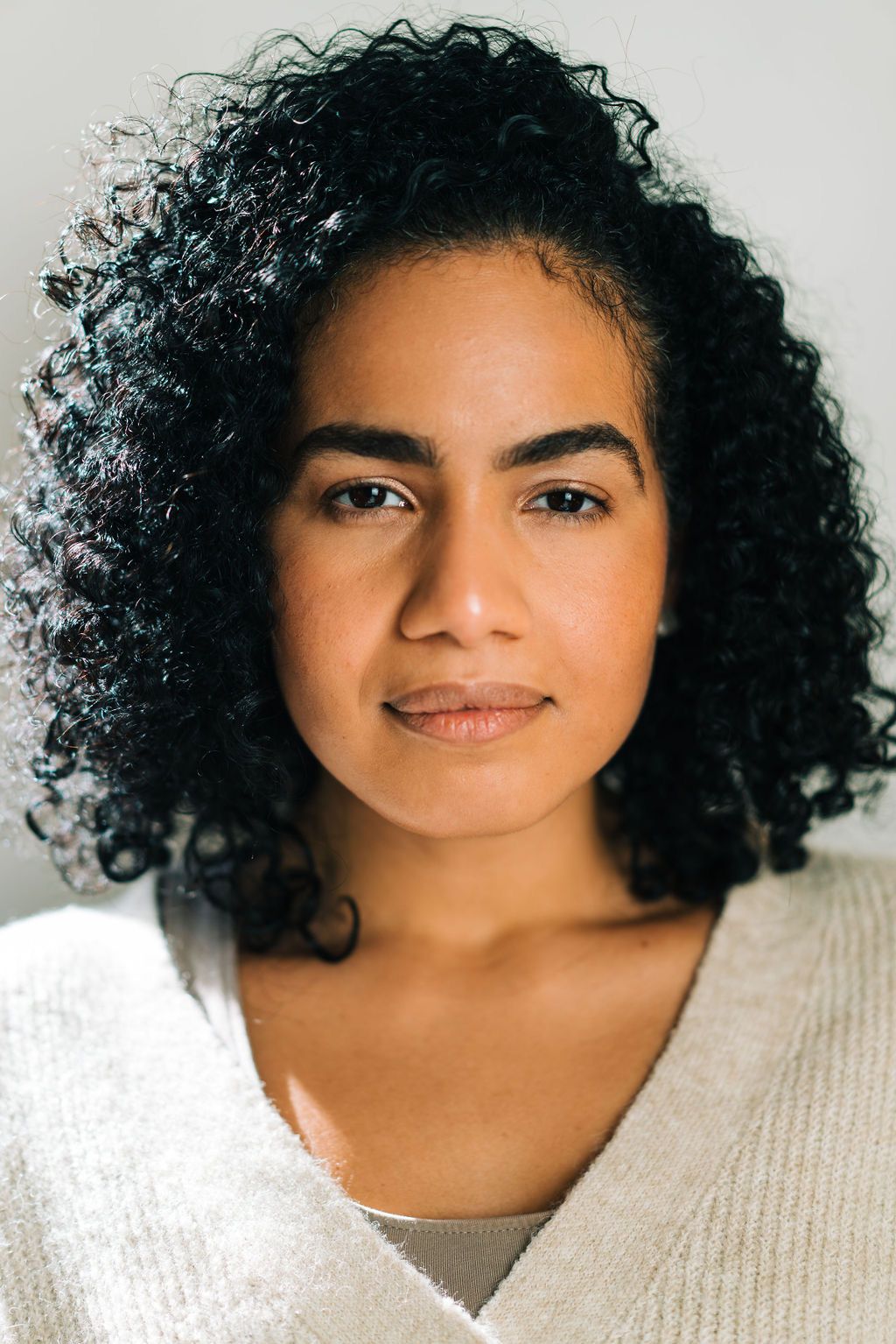 Portrait of a person with natural curly dark hair wearing a white sweater against a light background.